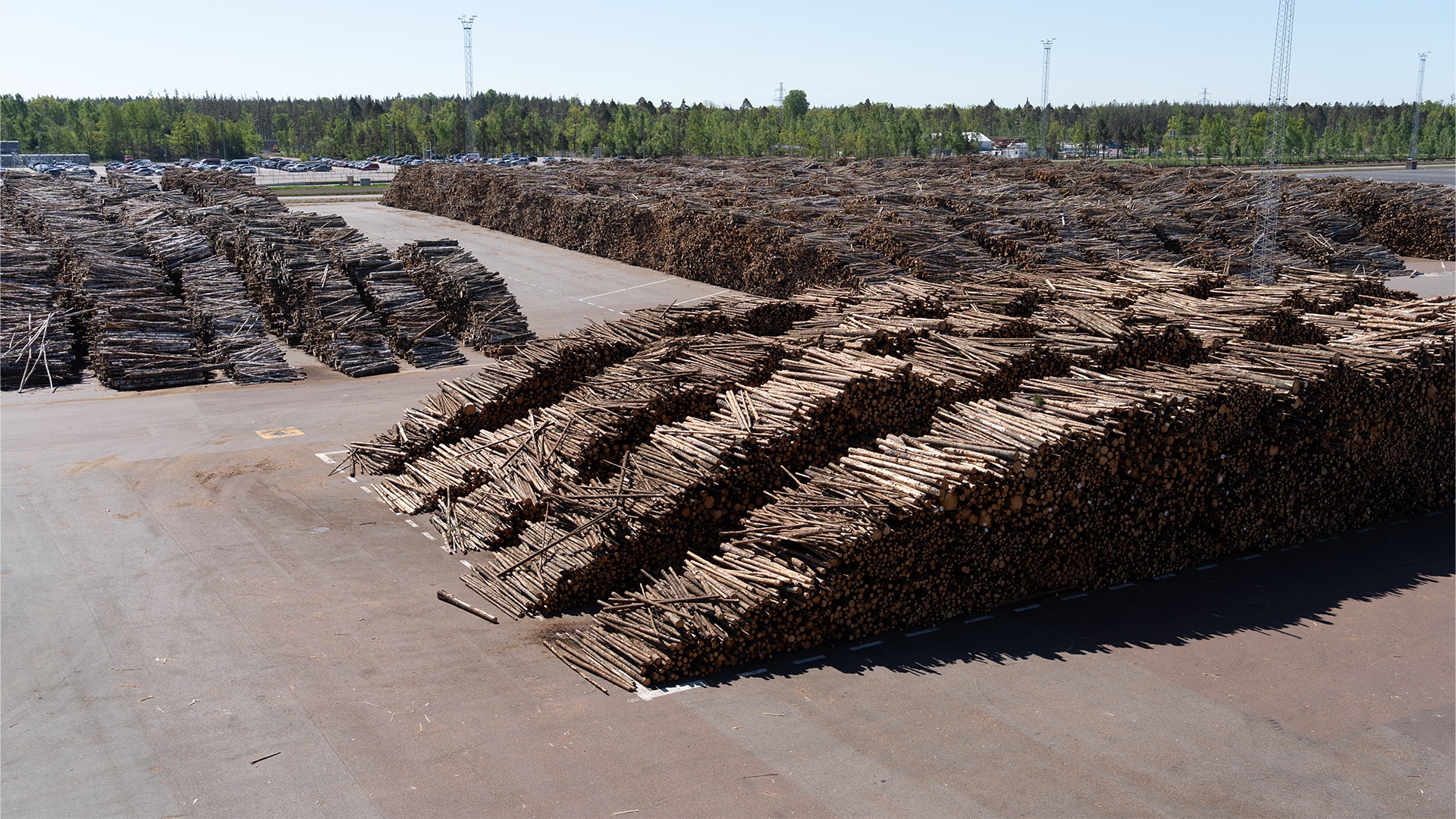 Stort lager med högar av trävara och stockar på en öppen betongyta. Skog och industrifacilitet syns i bakgrunden.