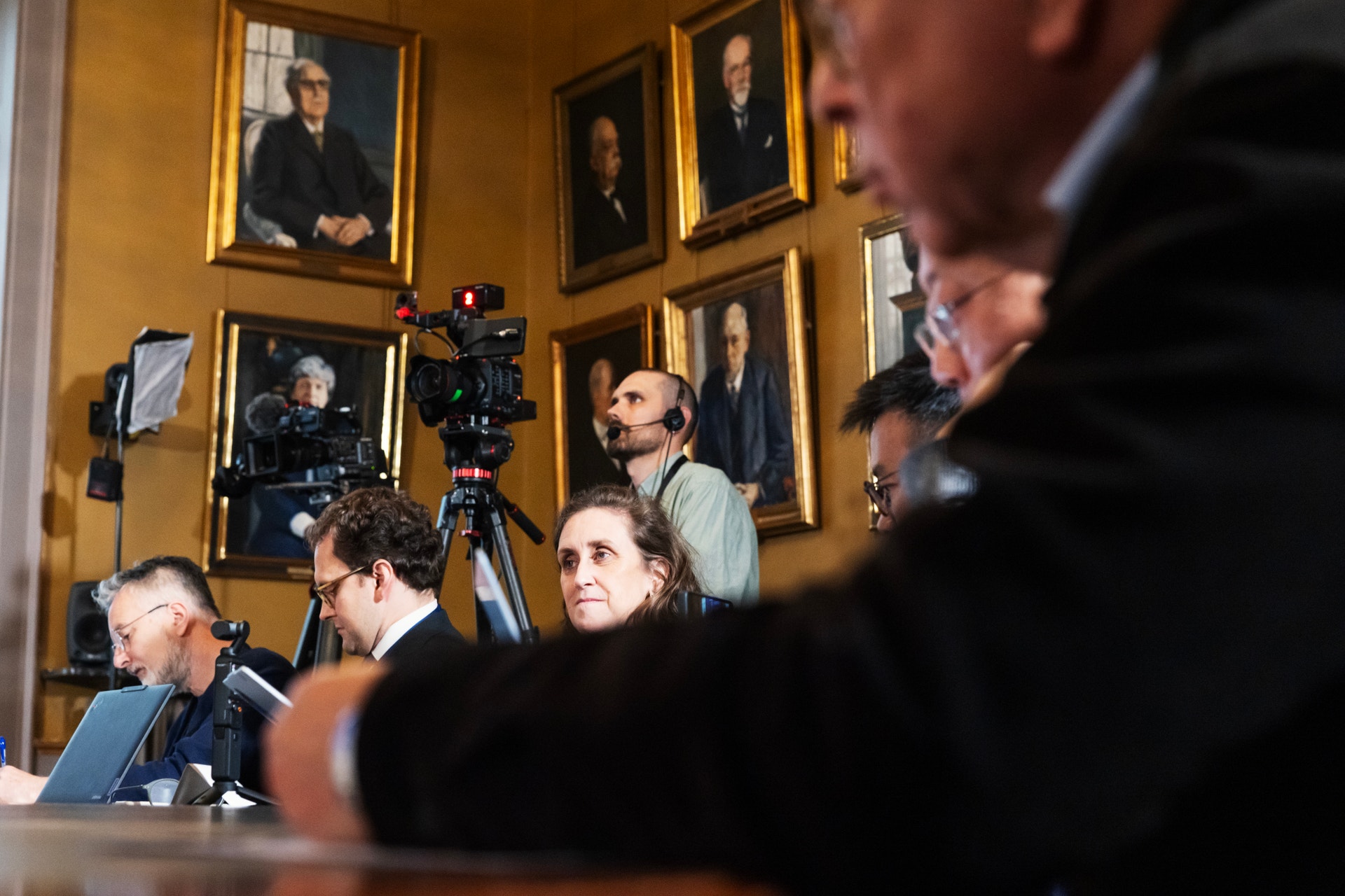 Journalister och en filmkamera i ett rum med inramade porträttmålningar på väggen, under en presskonferens.