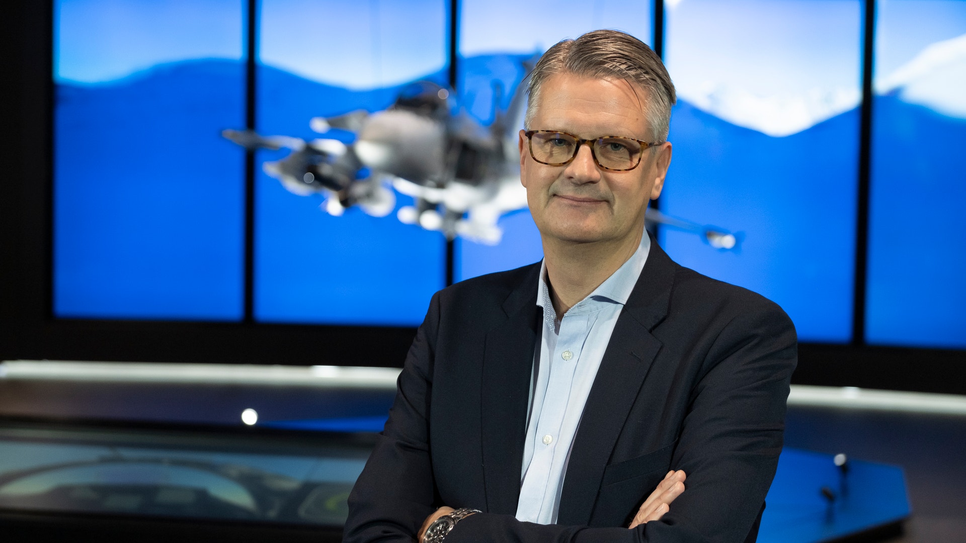 Man i kostym och glasögon står framför en skärm som visar ett flygplan mot blå himmel och berg.
