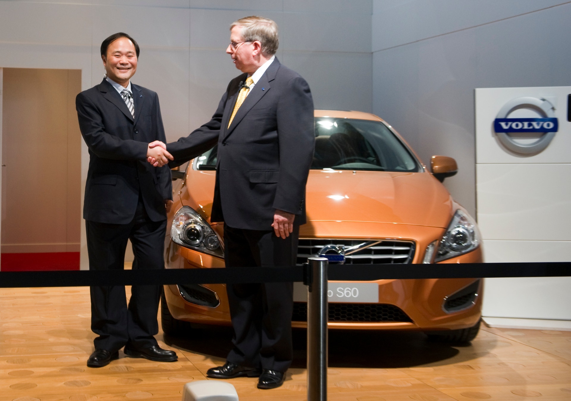 Två män i kostym skakar hand framför en orangefärgad Volvo S60 på en utställning.