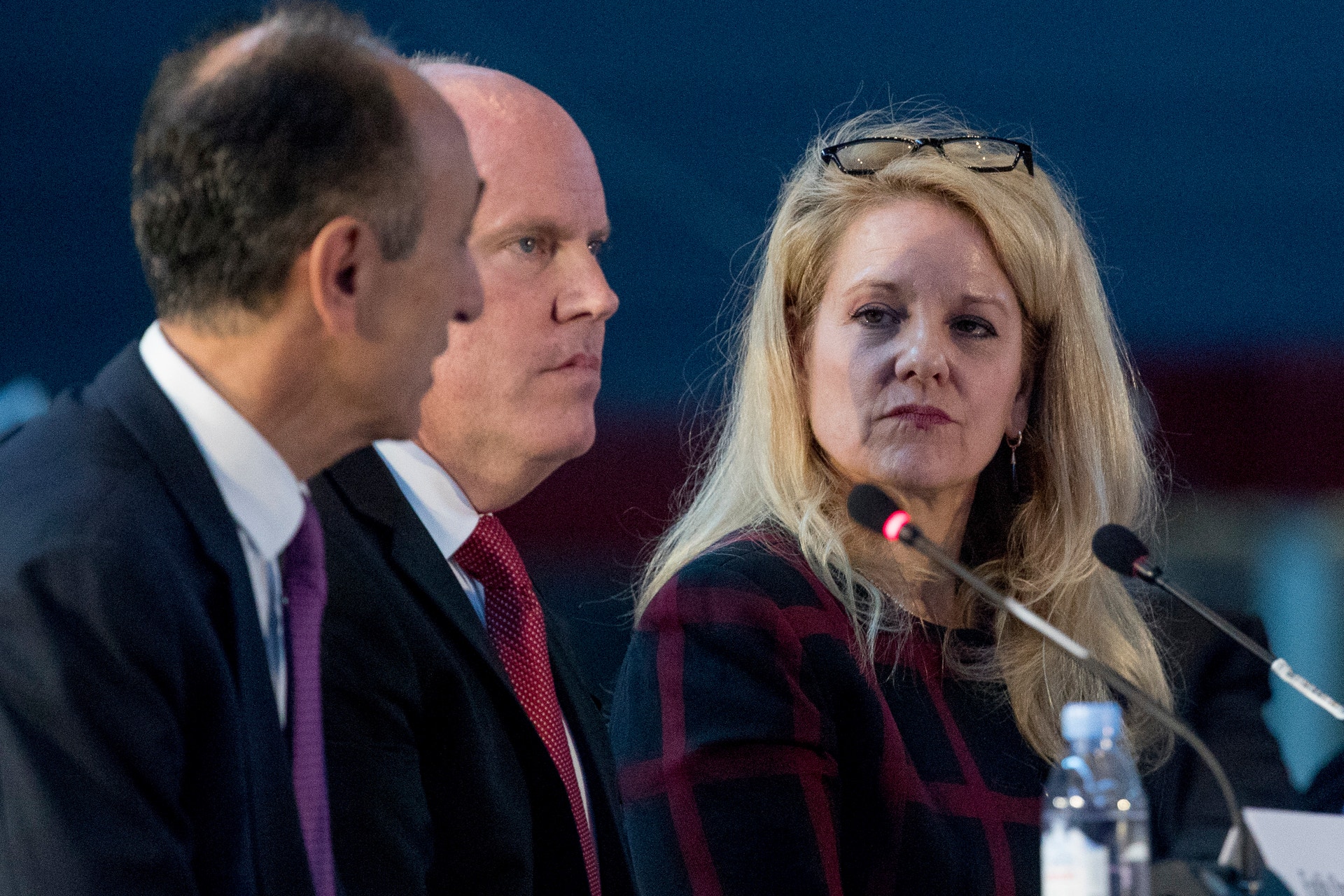 Tre personer i formell klädsel sitter vid ett bord med mikrofoner framför sig under en konferens eller paneldiskussion.