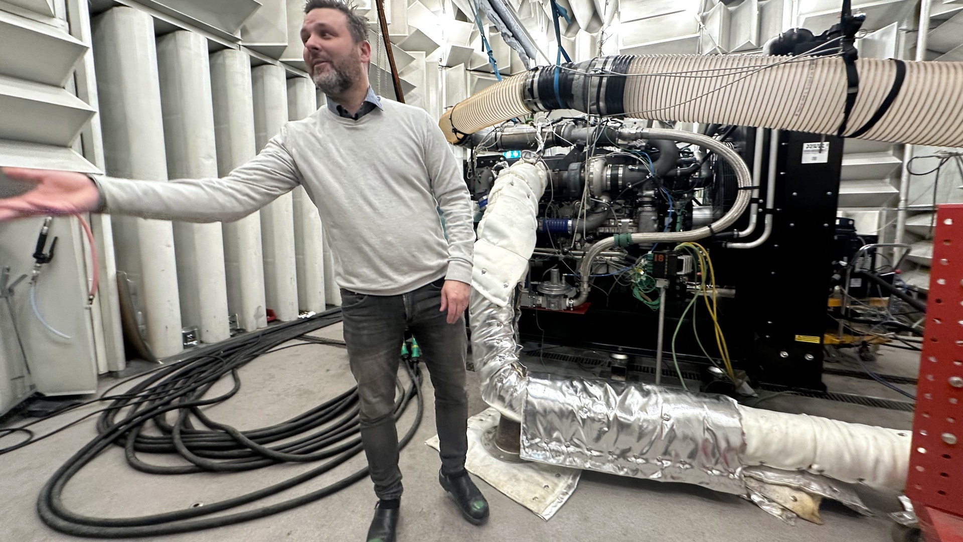 En man presenterar en stor industrimotor som är delvis lindad i vit isoleringsmaterial i en fabriksmiljö.