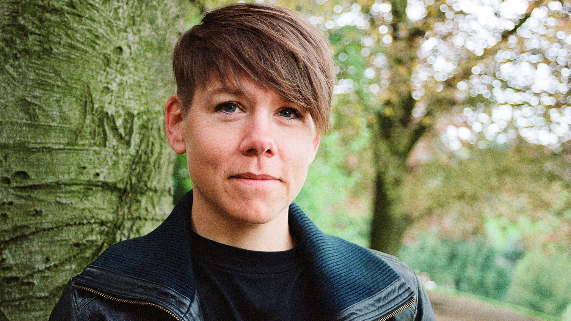 Porträtt av en ung kvinna med kort brunt hår, klädd i svart polotröja och mörkgrå jacka, lutar sig mot en stor trädstam i en grön naturmiljö med solljus som filtreras genom löven.