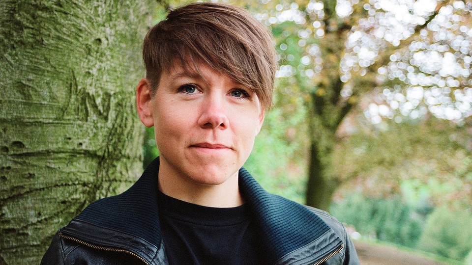 Porträtt av en ung kvinna med kort brunt hår, klädd i svart polotröja och mörkgrå jacka, lutar sig mot en stor trädstam i en grön naturmiljö med solljus som filtreras genom löven.