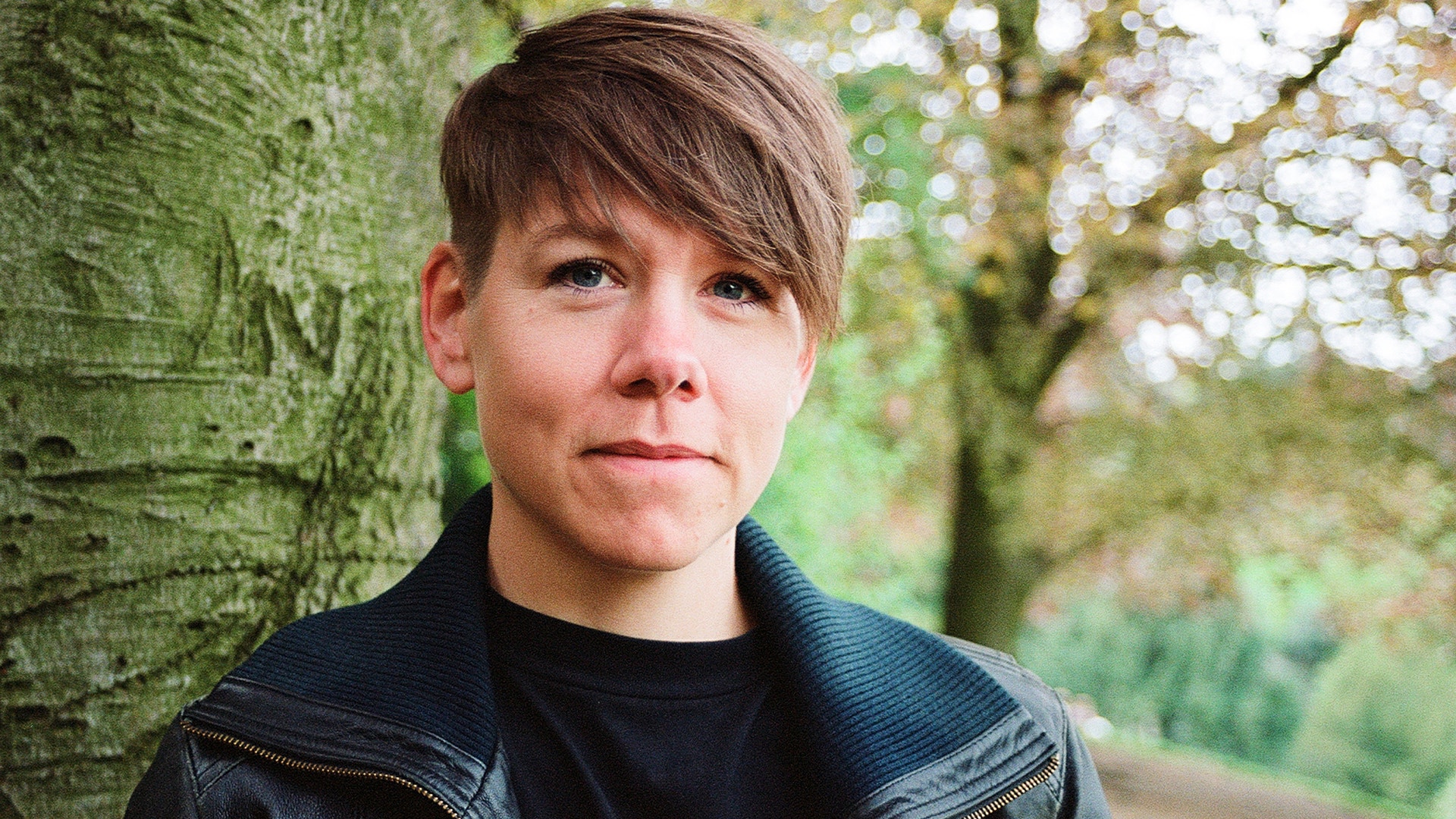 Porträtt av en ung kvinna med kort brunt hår, klädd i svart polotröja och mörkgrå jacka, lutar sig mot en stor trädstam i en grön naturmiljö med solljus som filtreras genom löven.