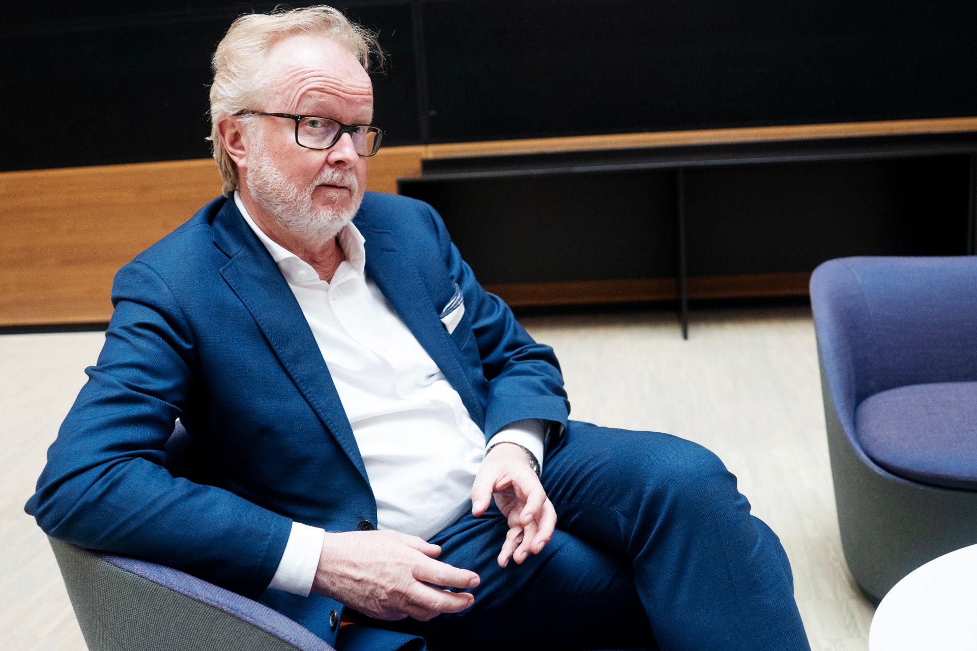 En man i blå kostym sitter i en modern kontorsmiljö och tittar åt sidan.