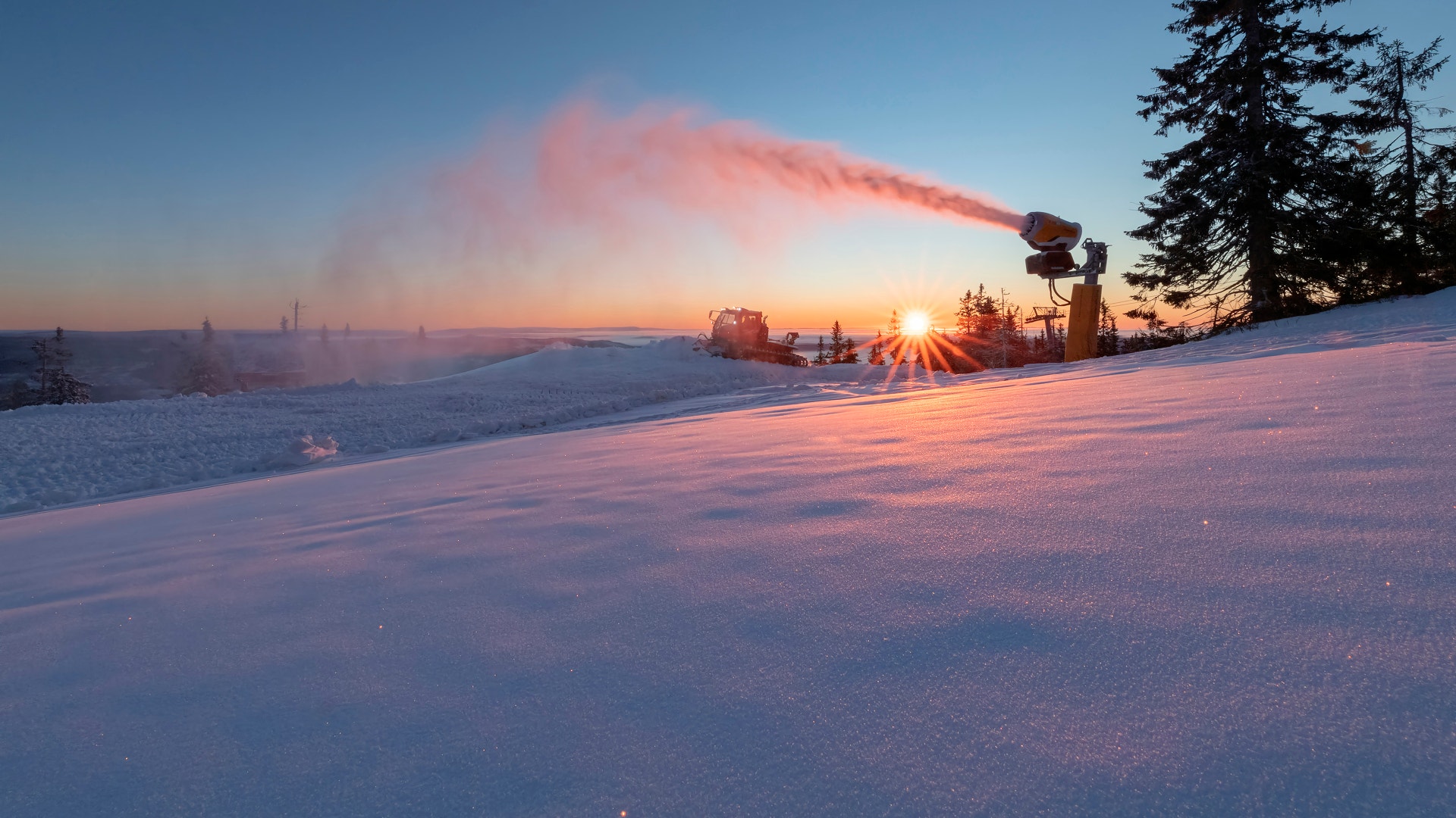 En snökanon sprutar snö i en skidbacke i solnedgången, med snötäckta granar och en lysande sol vid horisonten.