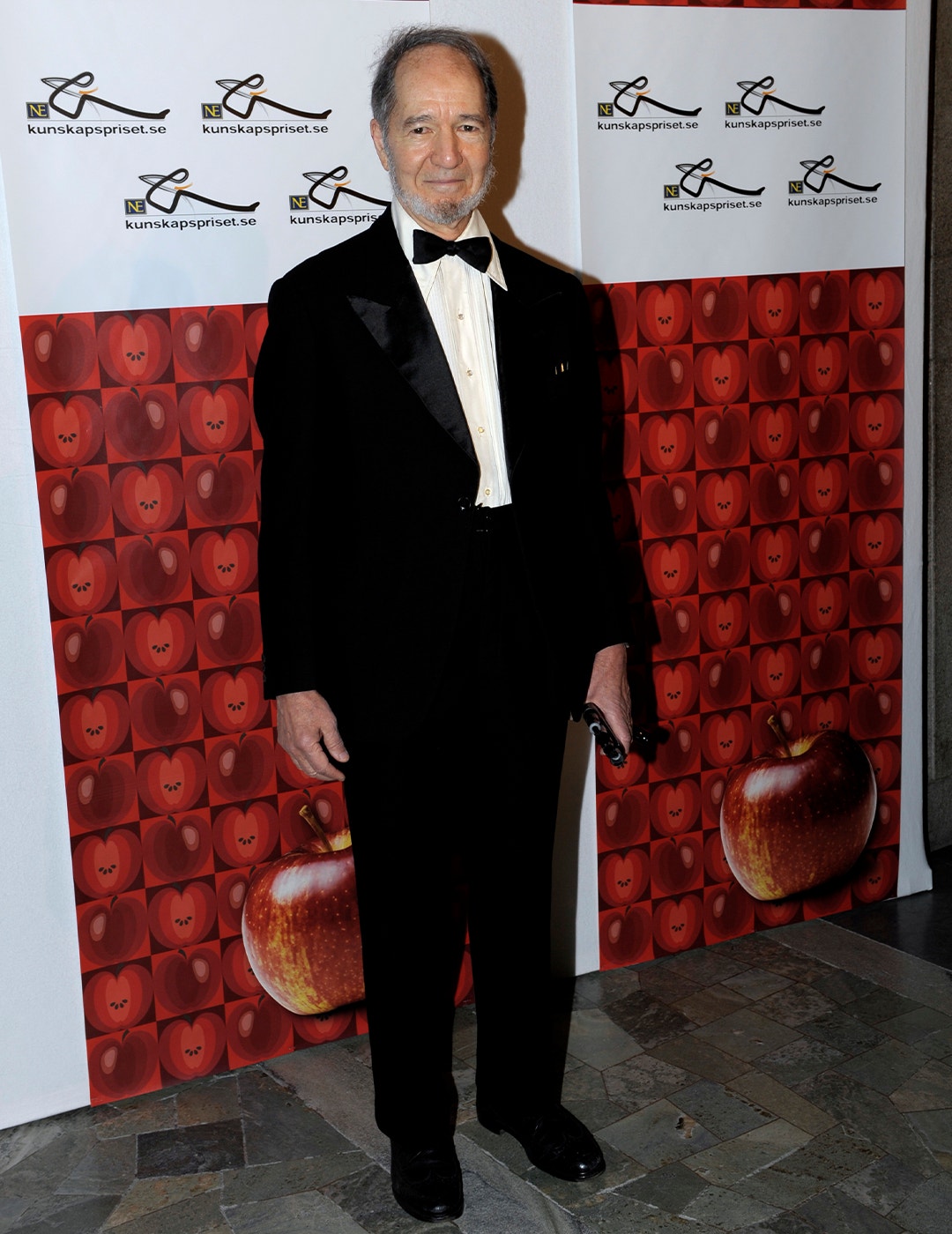 Äldre man i smoking och fluga framför röd bakgrund med äppelmotiv vid formell event.