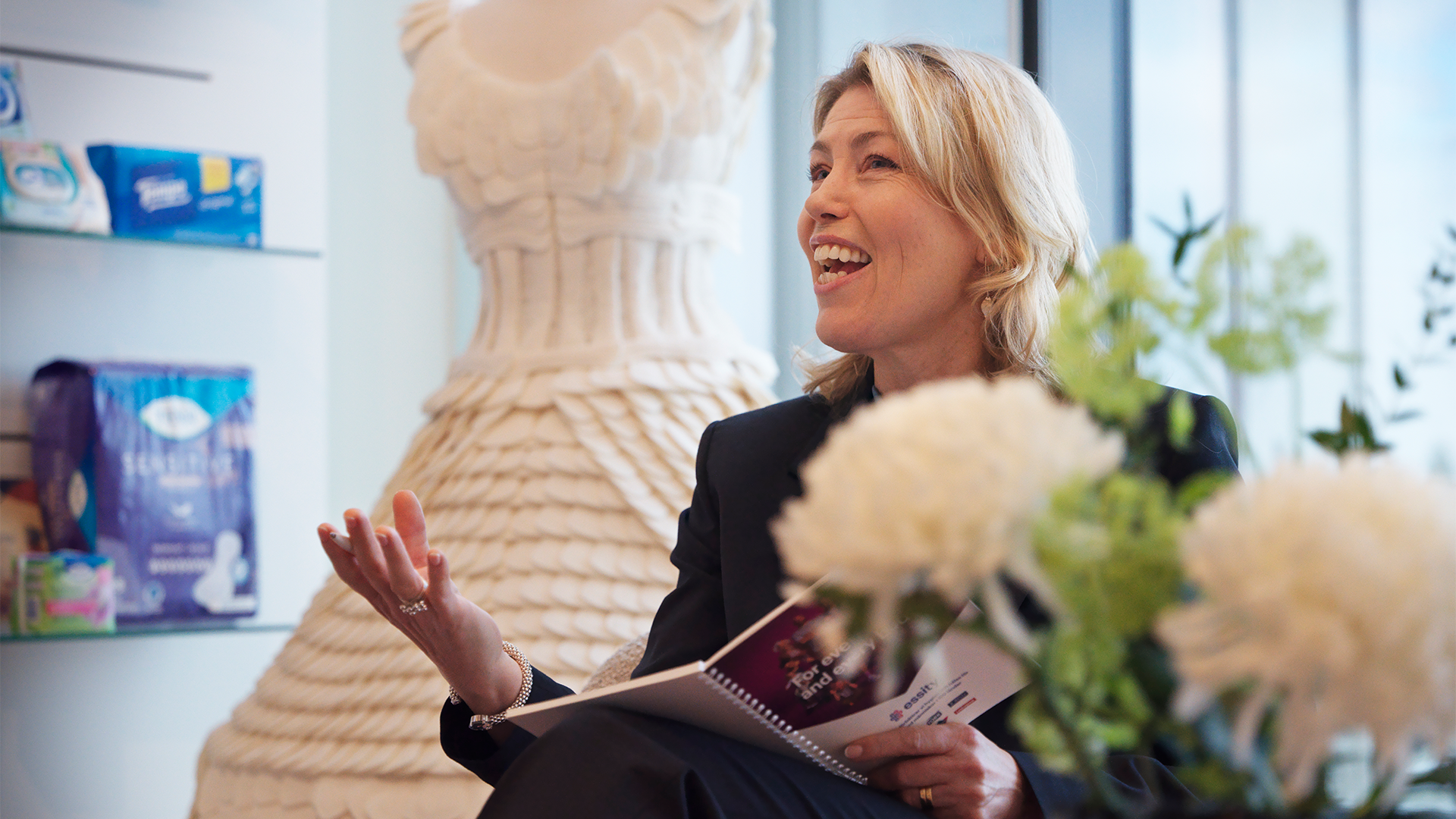 Kvinna i svart jacka håller en skrivplatta och presenterar med ett leende i ett modernt kontor med hyllor, växter och dekorativa föremål i bakgrunden.