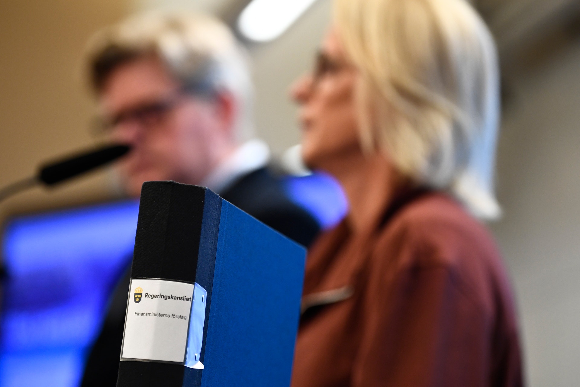 En mikrofon med blått skydd i förgrunden under en presentation. I bakgrunden ses två personer som presenterar.