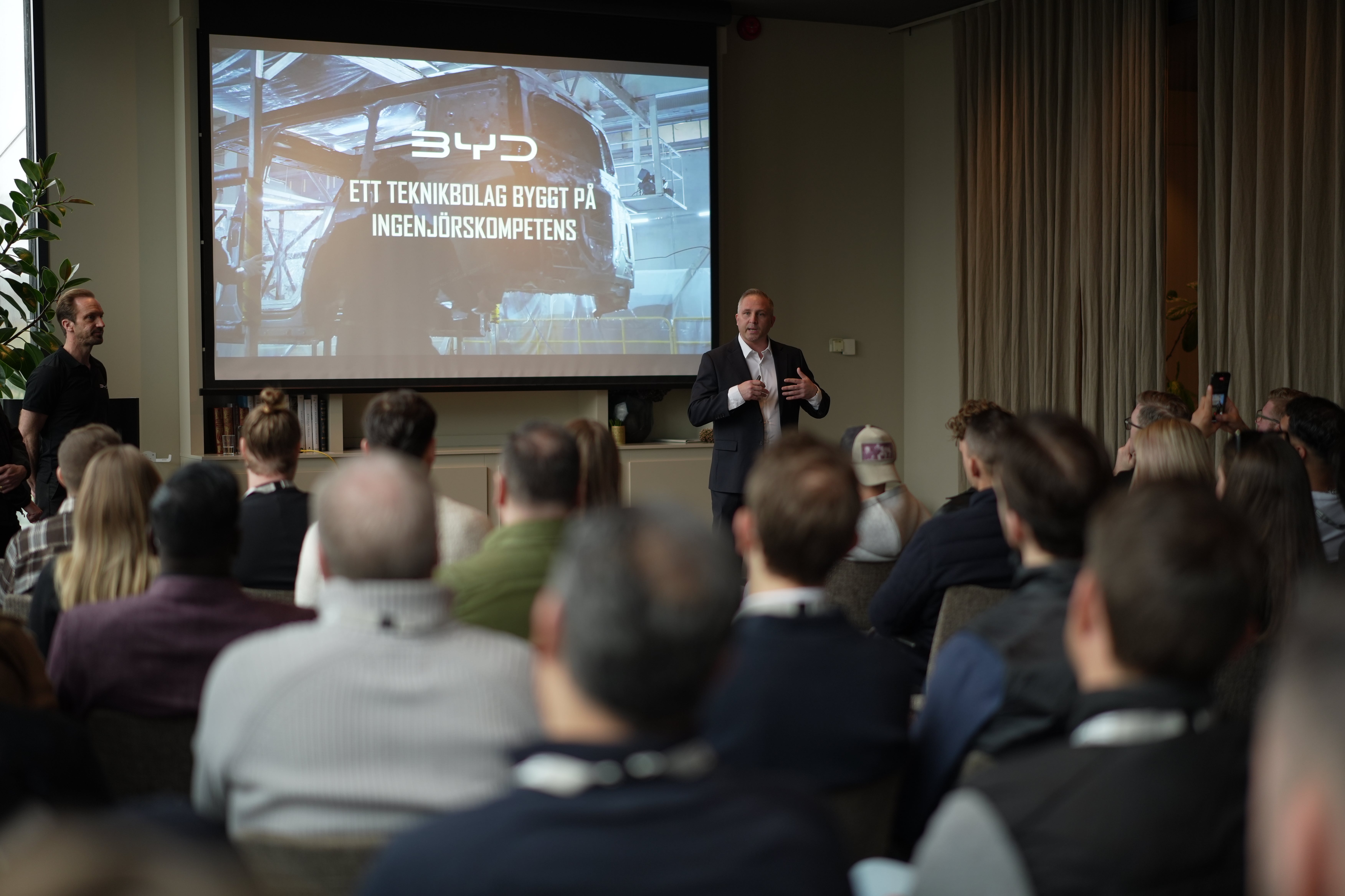 En presentatör håller ett föredrag inför en publik i ett konferensrum med en projektionsskärm i bakgrunden.