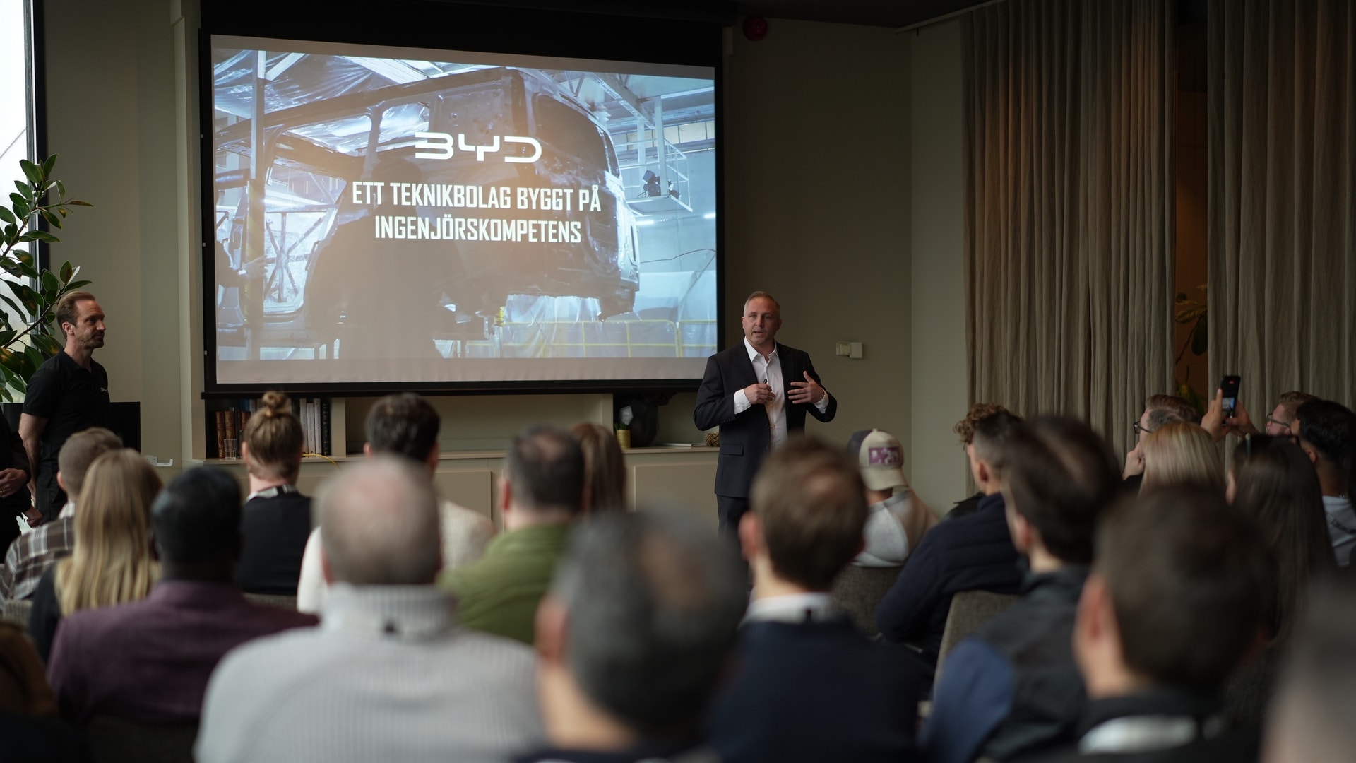 En presentatör håller ett föredrag inför en publik i ett konferensrum med en projektionsskärm i bakgrunden.