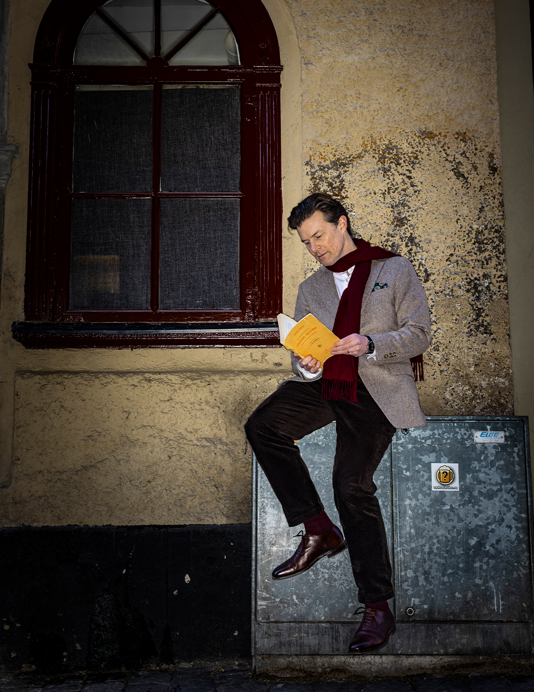 Man sitter på en metallåda mot en sliten vägg och läser en gul bok