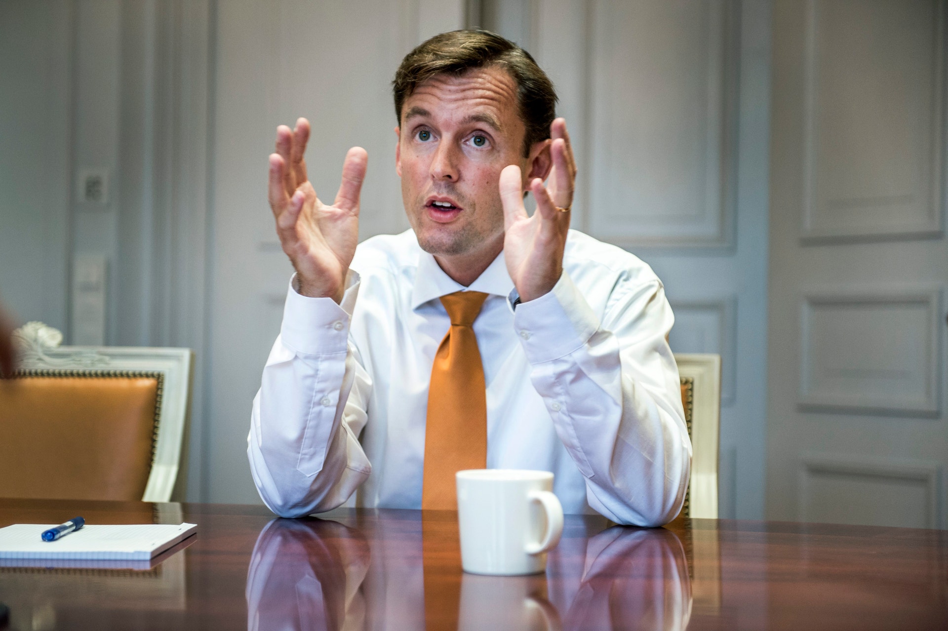 En man i vit skjorta och orange slips sitter vid ett bord och gestikulerar med båda händerna medan han pratar.