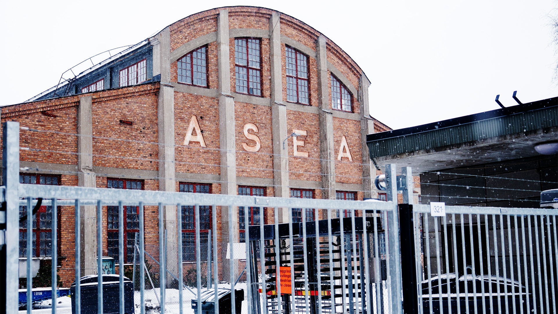 En historisk tegelbyggnad med texten "ASEA" på fasaden. Byggnaden har karakteristiska välvda fönster, vita betongpelare och en industriell arkitektur från början av 1900-talet. En vit metallstängsel omger byggnaden.