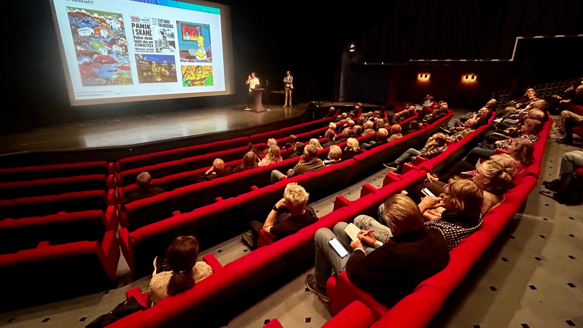 Publik sitter i röda biosäten och tittar på en presentation på en stor skärm i en biograf.