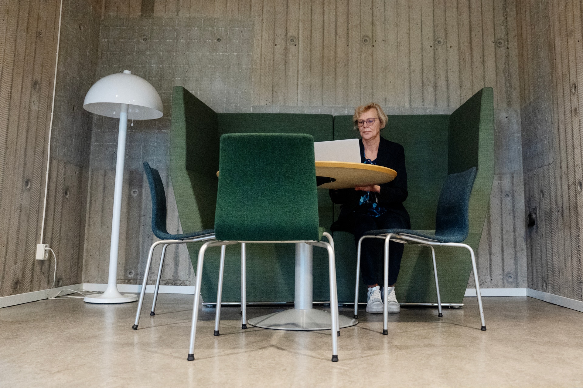 En kvinna med kort ljust hår och glasögon sitter vid ett runt bord med en laptop i en grön fåtölj med höga kanter, i ett rum med betongväggar.