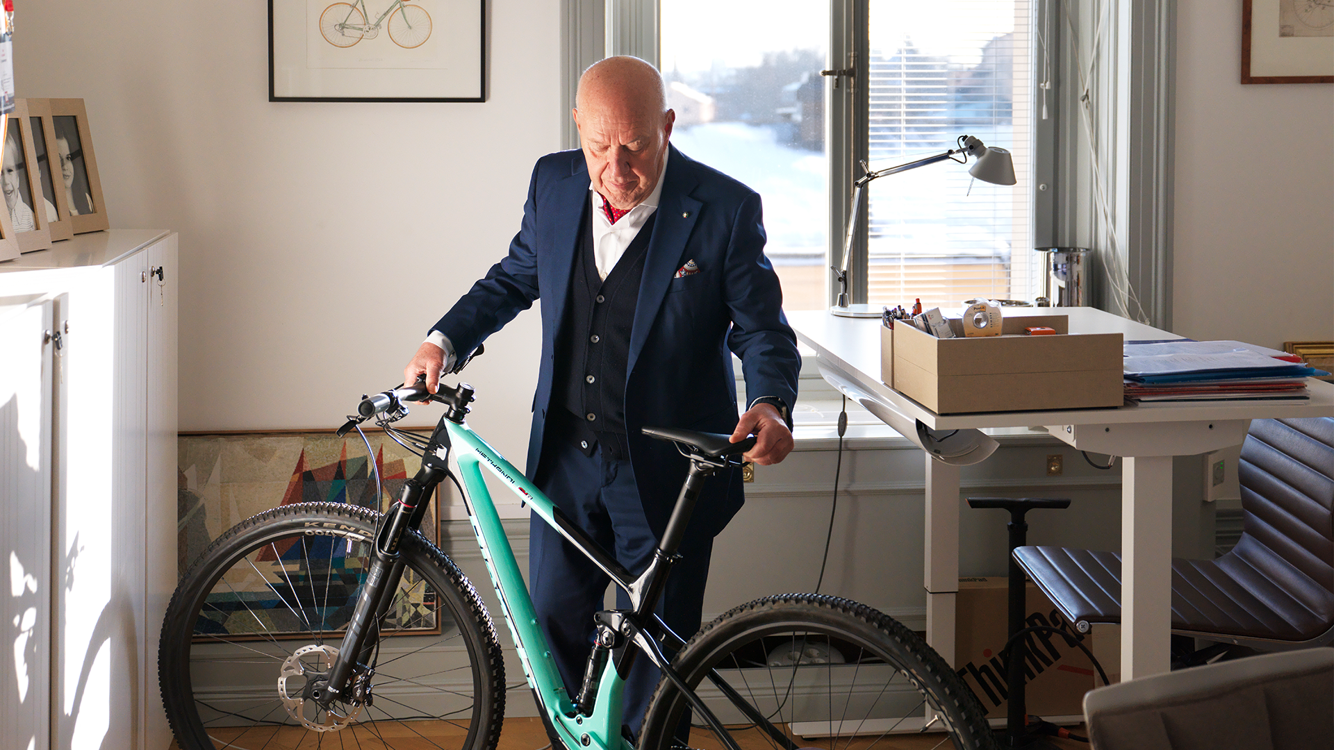 En man i mörkblå kostym står med en turkos cykel i ett modernt kontor med stora fönster.