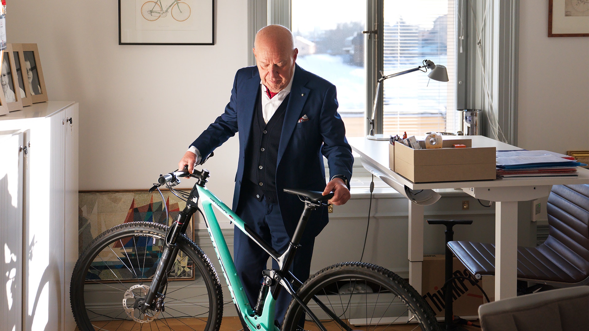En man i mörkblå kostym står med en turkos cykel i ett modernt kontor med stora fönster.