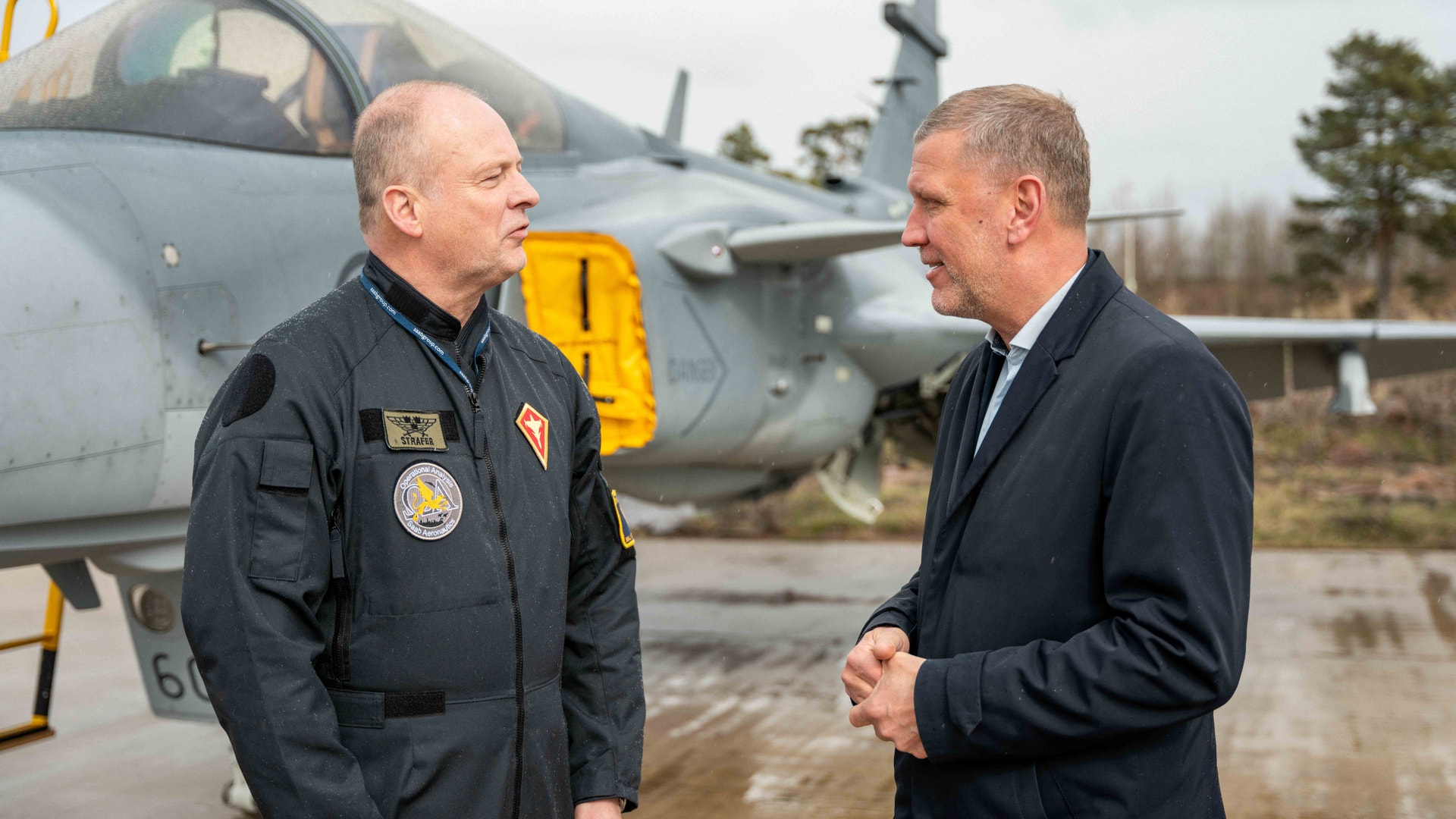En militär officer i uniform och en man i kostym samtalar framför ett militärflygplan på en flygbas.