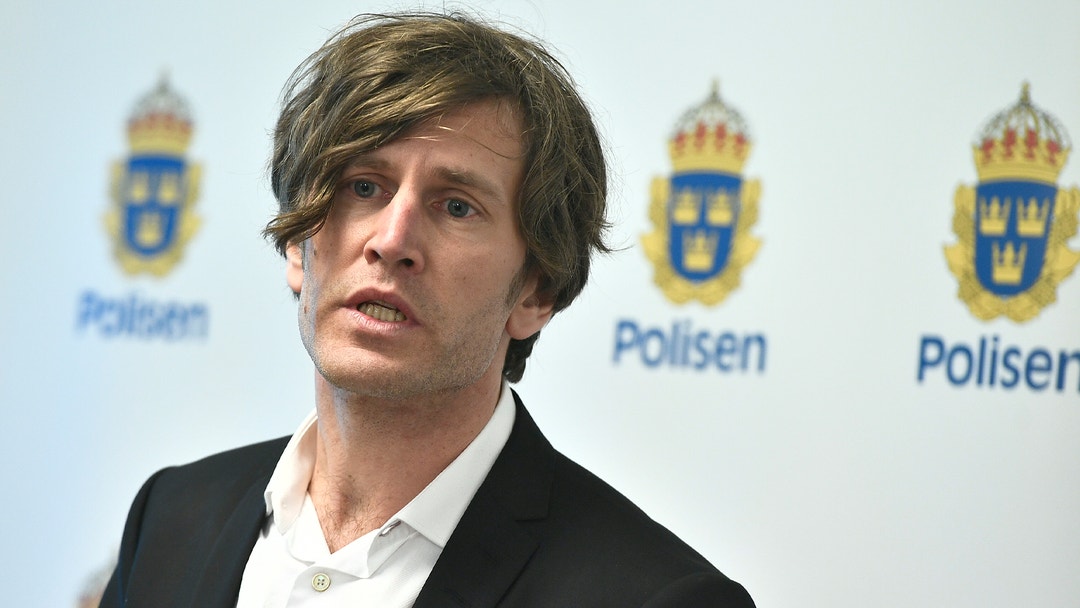 Man i mörk kavaj och vit skjorta talar framför Polisens logotyp.
