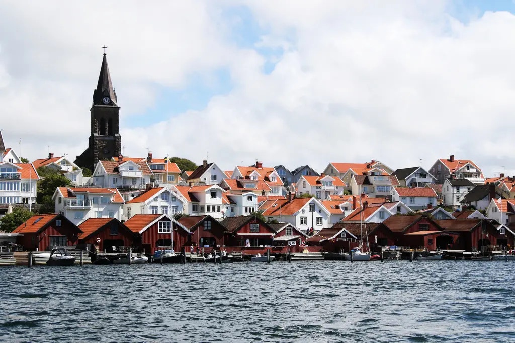 En kustnära by med vita hus med röda tak, traditionella röda båthus vid vattnet och en kyrka med torn i bakgrunden.