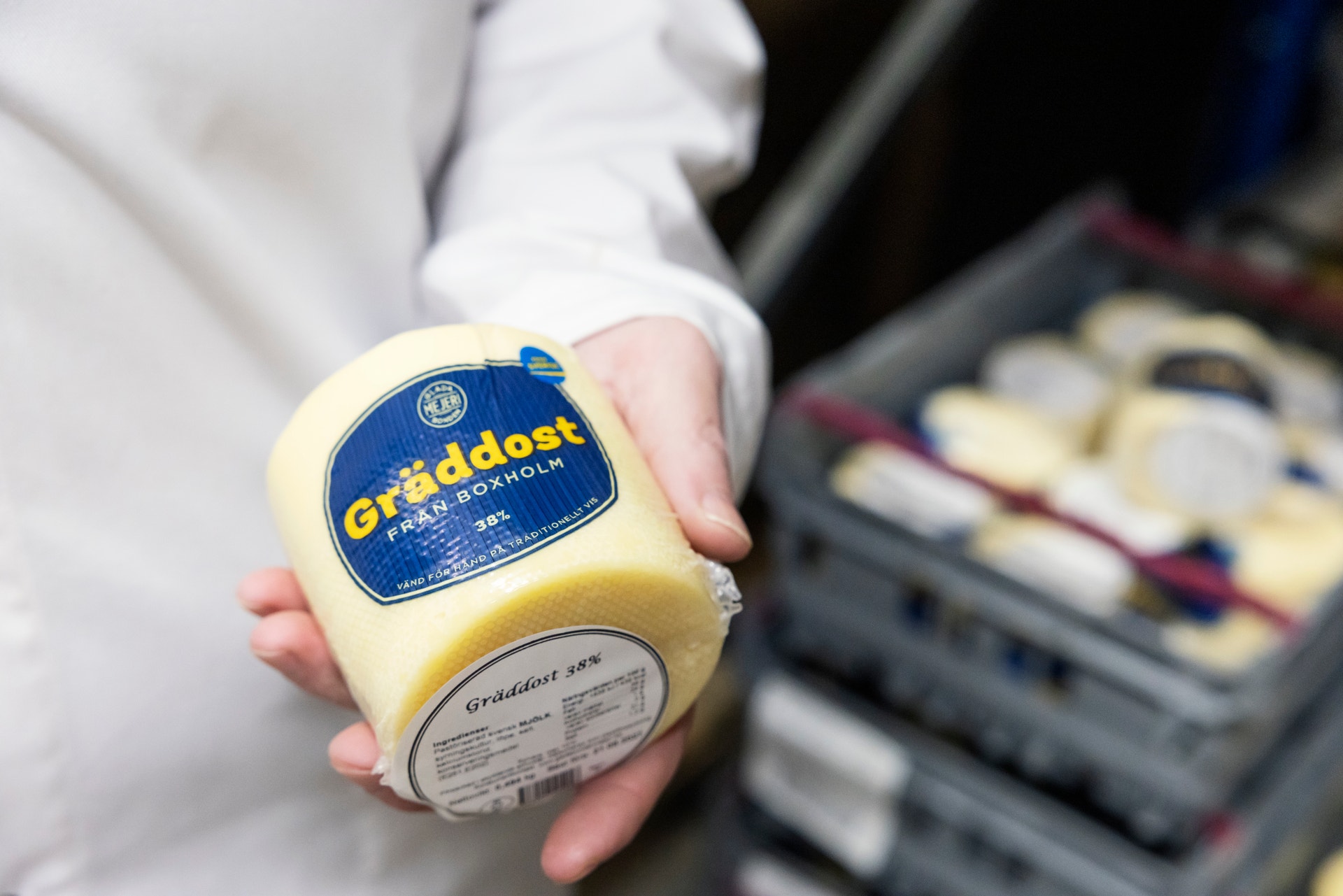 En hand håller en gul ostaprodukt med blå etikett från Glada bonden mejeri. I bakgrunden syns fler ostprodukter i en låda.