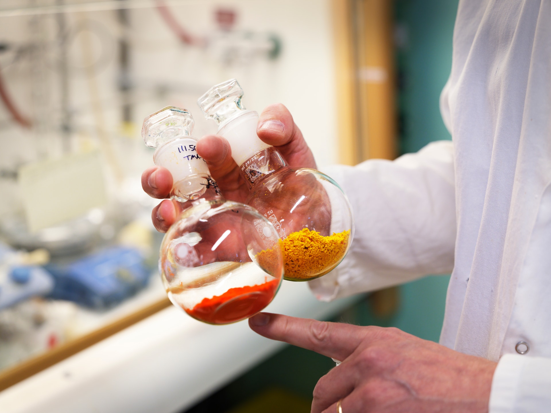 En person i vit laboratoriekittel håller en glaskolv med färgade kemiska vätskor - rött, gult och orange - i ett kemilaboratorium.
