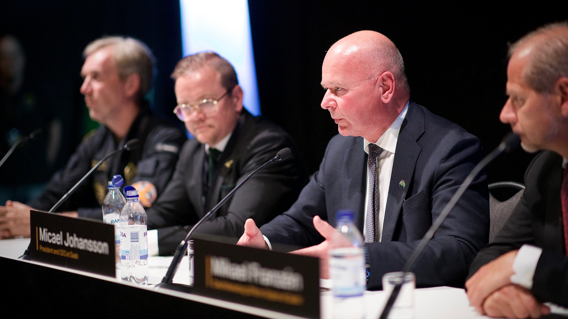 Fyra män i kostym sitter vid ett bord under en konferens eller paneldiskussion. De har mikrofoner och vattenflaskor framför sig.