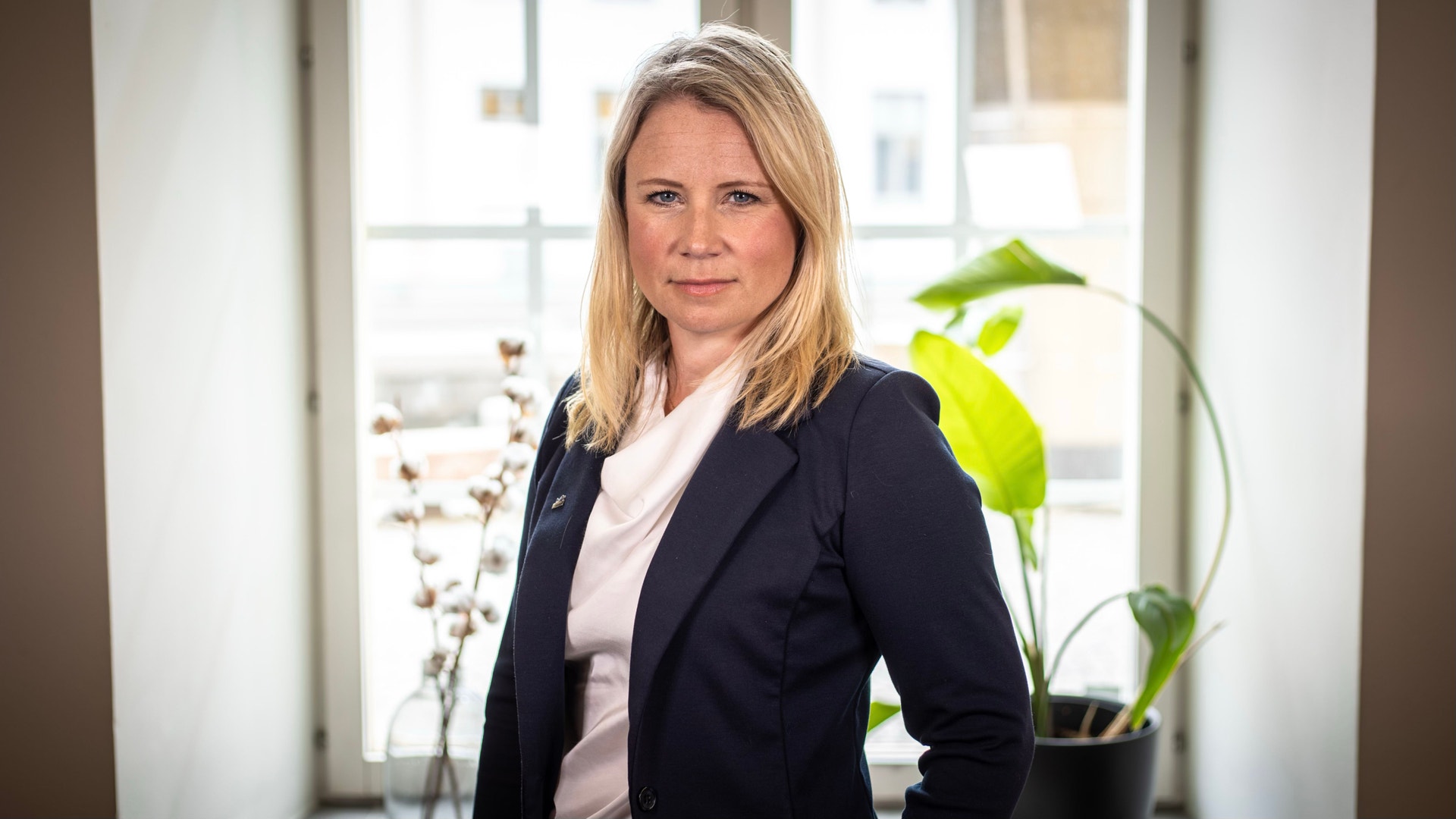 Porträttfoto av en kvinna i mörkblå kavaj och vit skjorta, fotograferad inomhus med växter i bakgrunden.