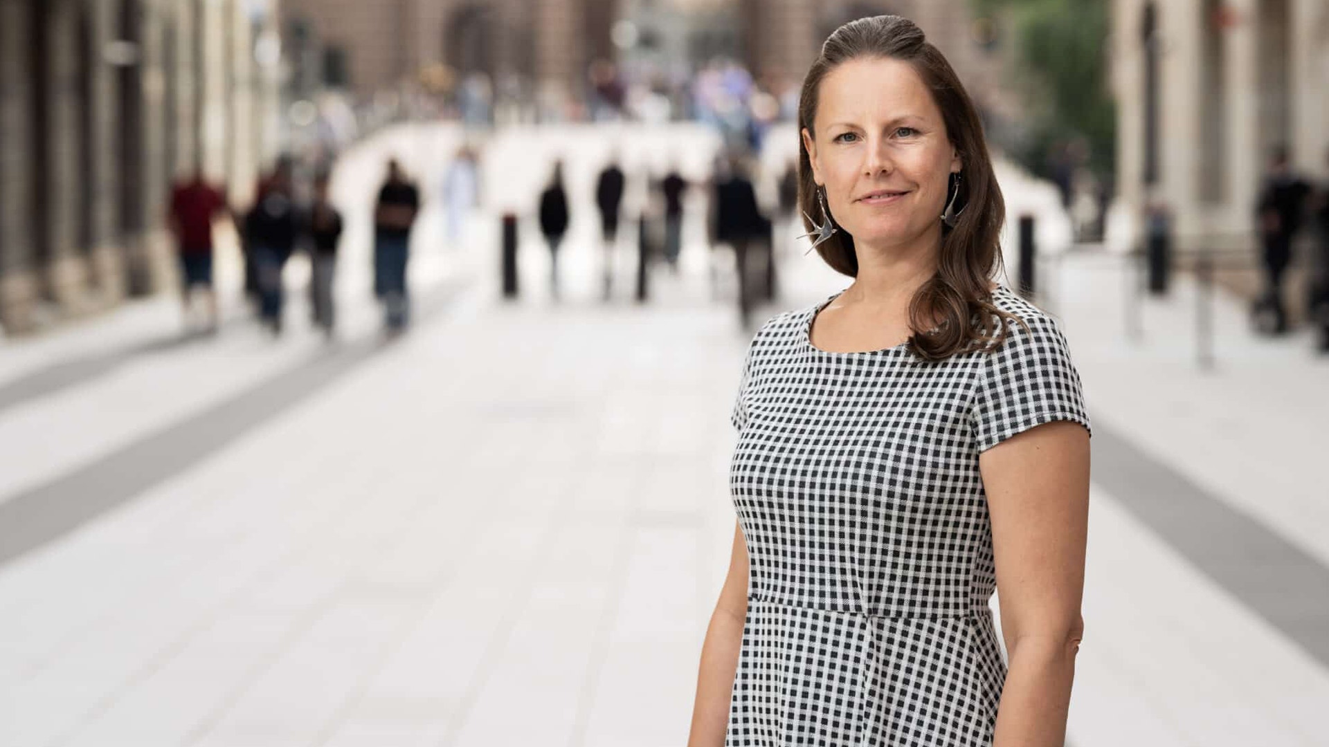 Porträttfoto av en kvinna i rutig klänning som står på en stadsgatа med människor i bakgrunden.