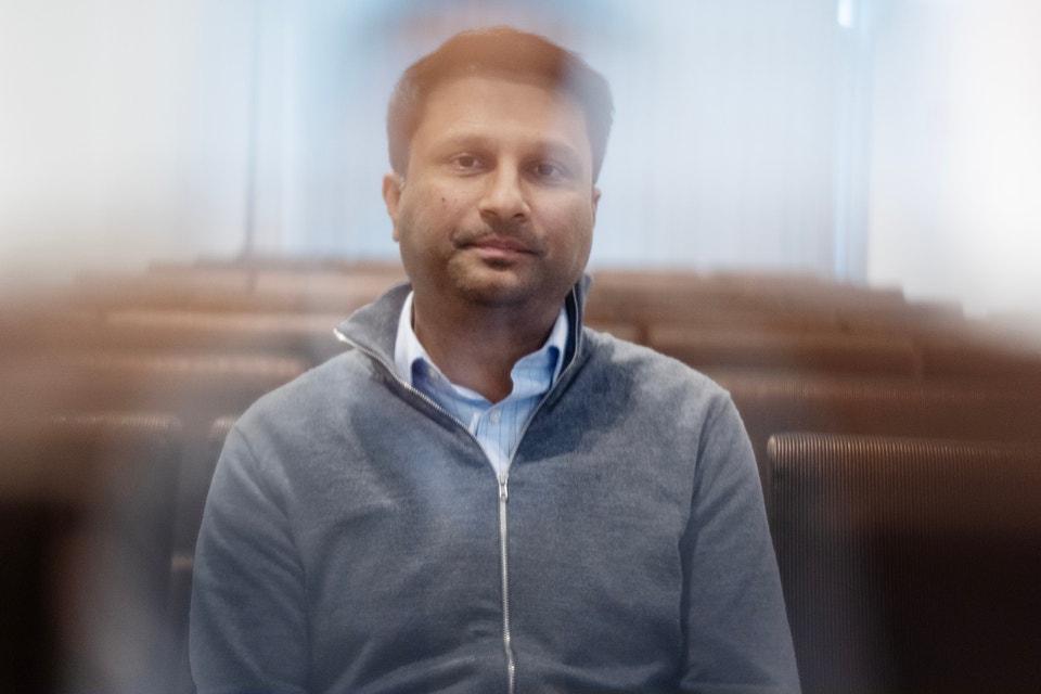 Porträtt av en man med kort brunt hår och skägg, klädd i en grå dragkedjetröja över en ljusblå skjorta. Han är fotograferad inomhus mot en suddig bakgrund med möbler, och tittar direkt mot kameran med ett lugnt uttryck.