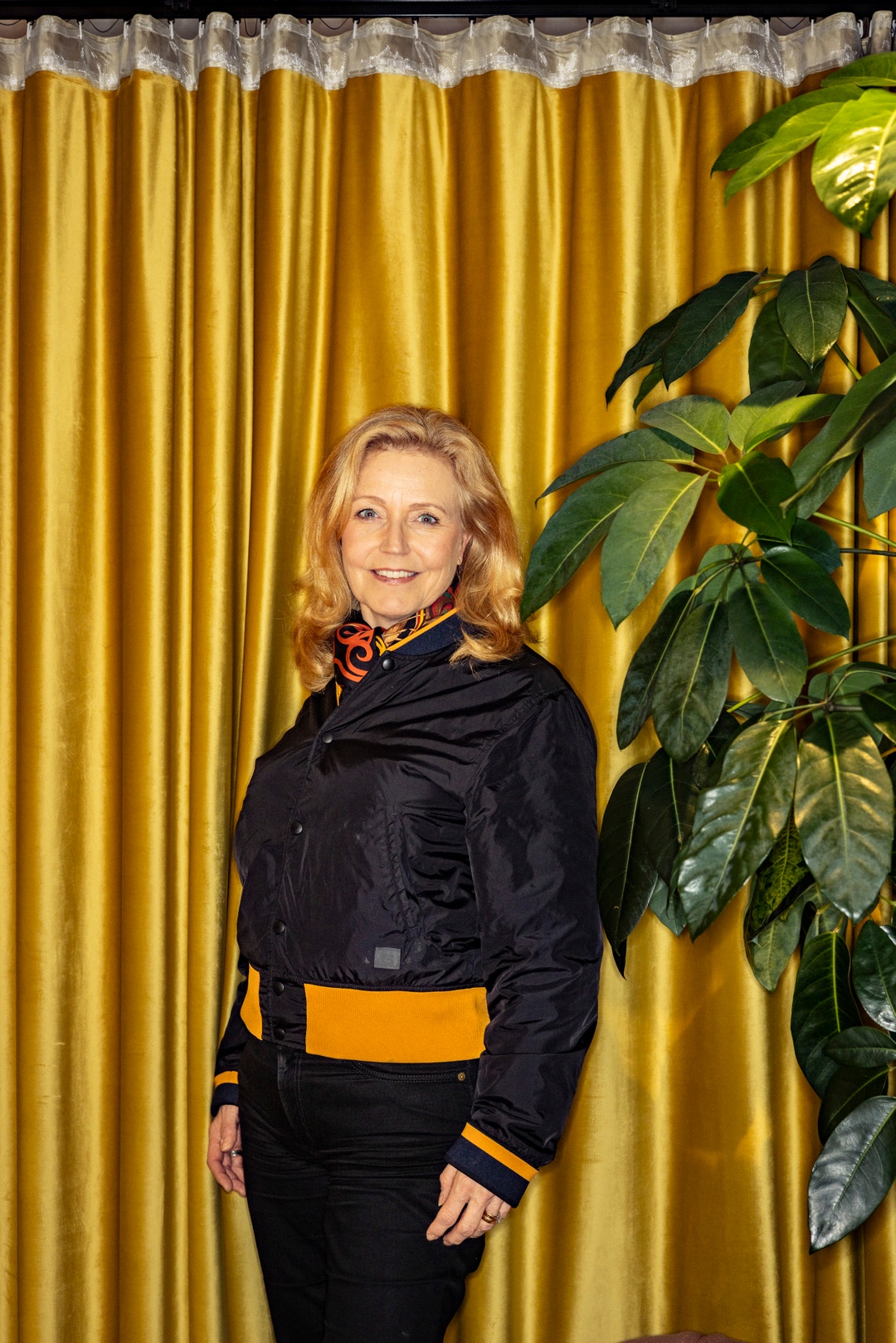 Porträtt av en kvinna som står framför en gul gardin med en grön växt bredvid sig.