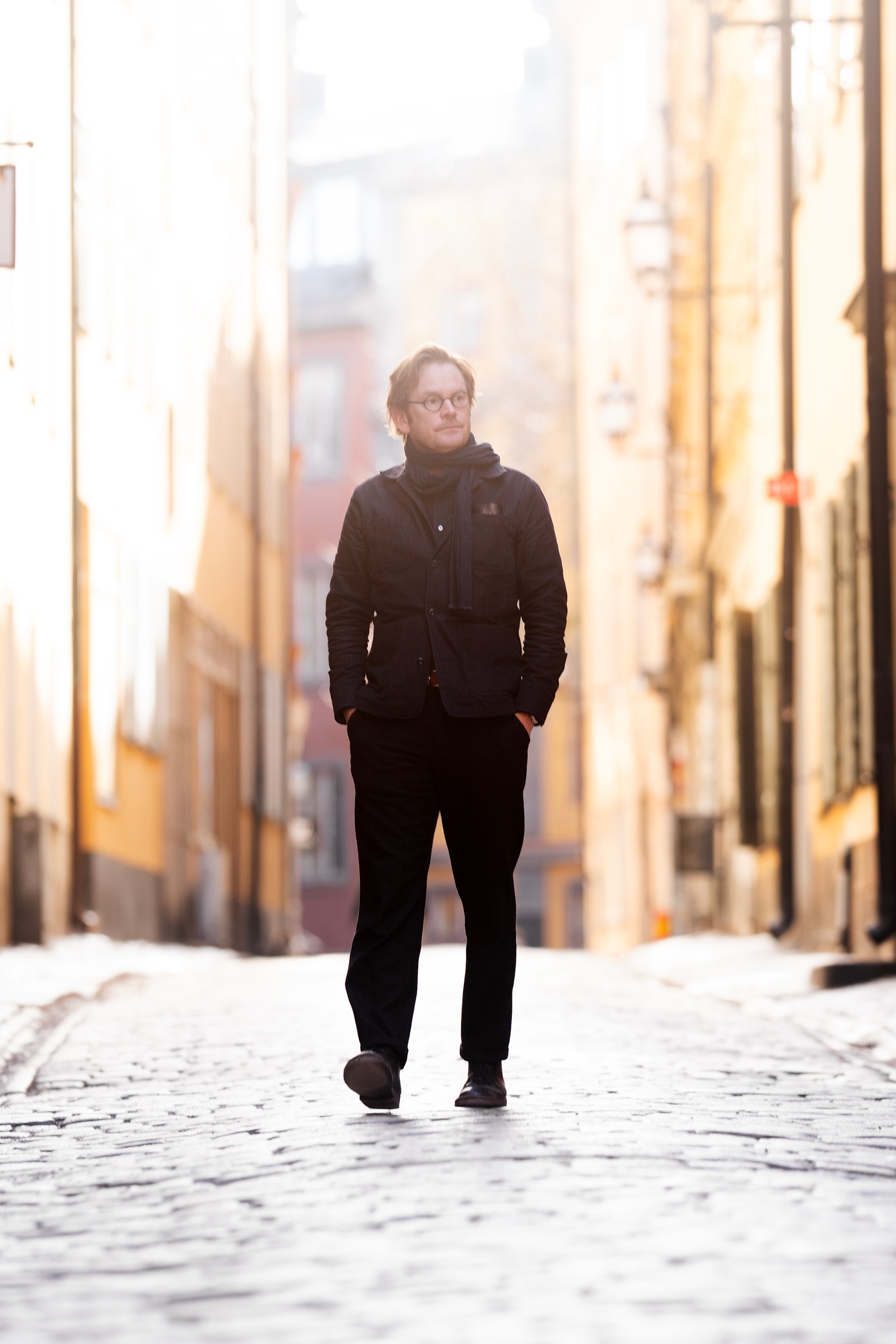 En person i svart jacka och byxor står i en smal, kullerstenlagd gata omgiven av gamla byggnader.