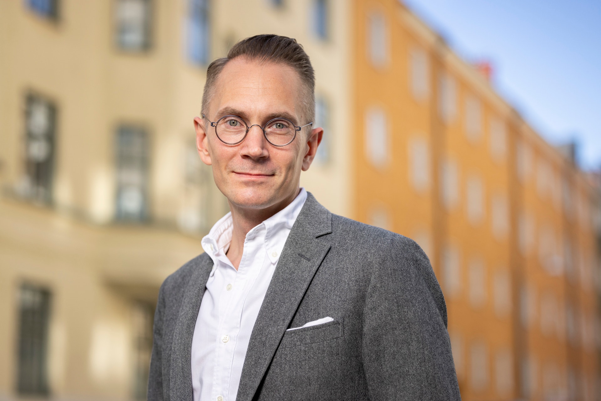 Porträtt av en man med glasögon och grå kavaj framför färgglada byggnader.