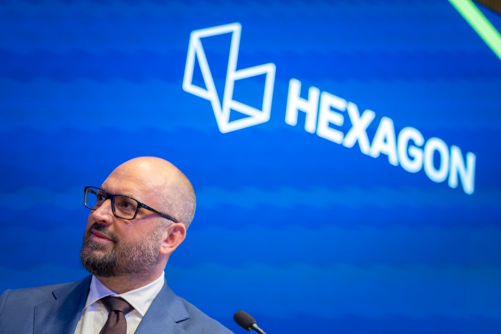 Man med glasögon och skägg talar vid en mikrofon framför en blå bakgrund med Hexagon-logotypen., 