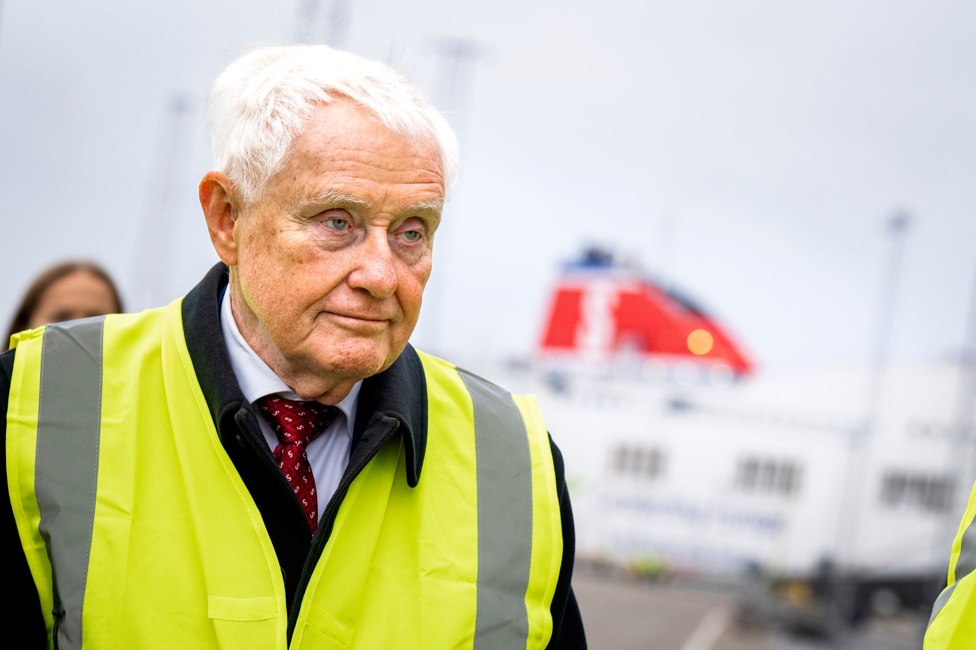 Äldre man med vitt hår i gul reflexväst står utomhus i en hamn. I bakgrunden syns en stor färja.
