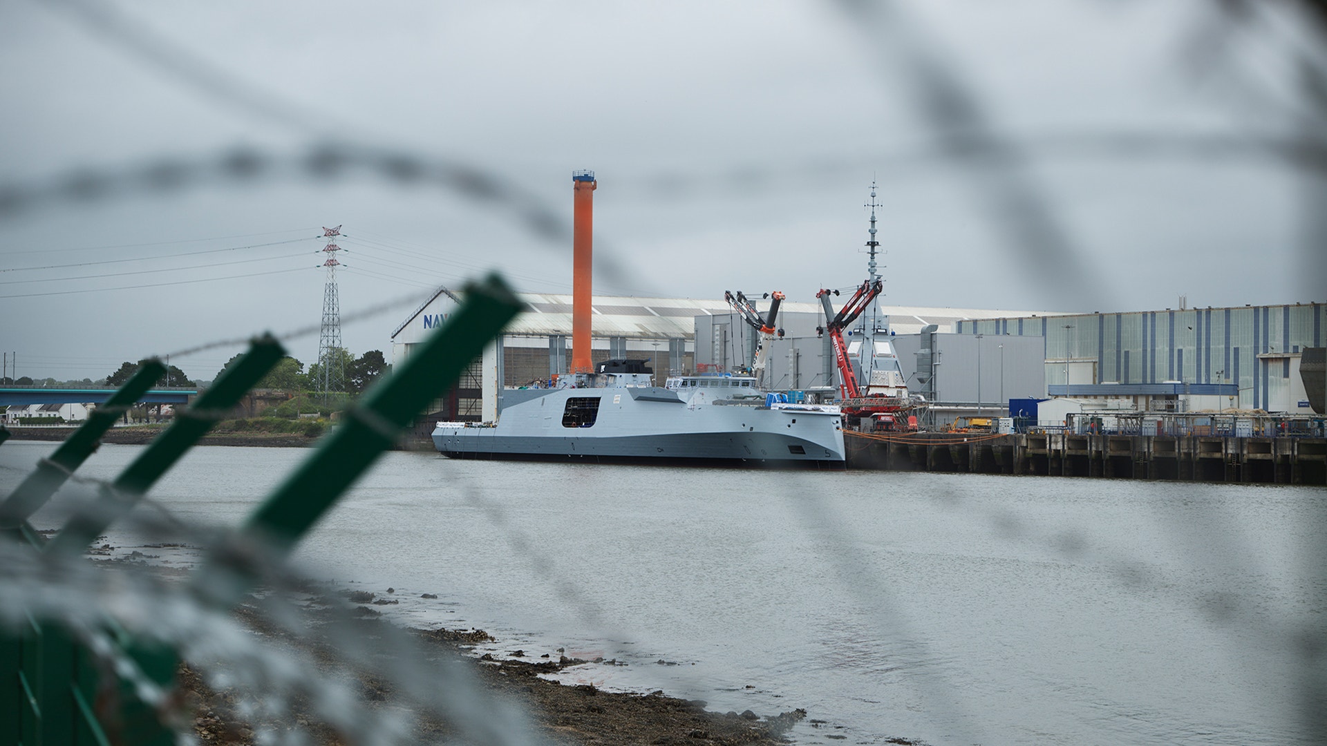 En vit och blå fregatt förtöjd vid en industriell hamn med fabriker och skorstenar i bakgrunden.