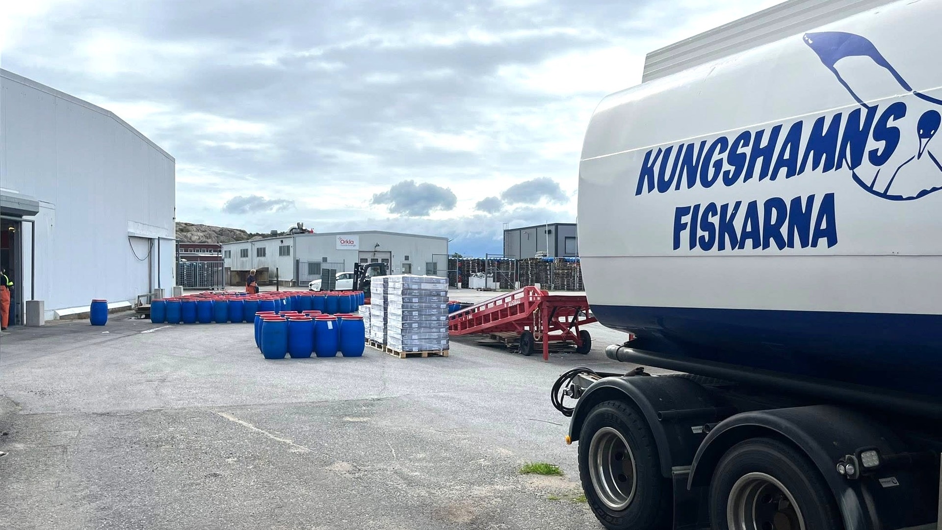 En lastbil märkt Kungshamsfiskarna står parkerad på en industriell gård med byggnader och blå behållare i bakgrunden.