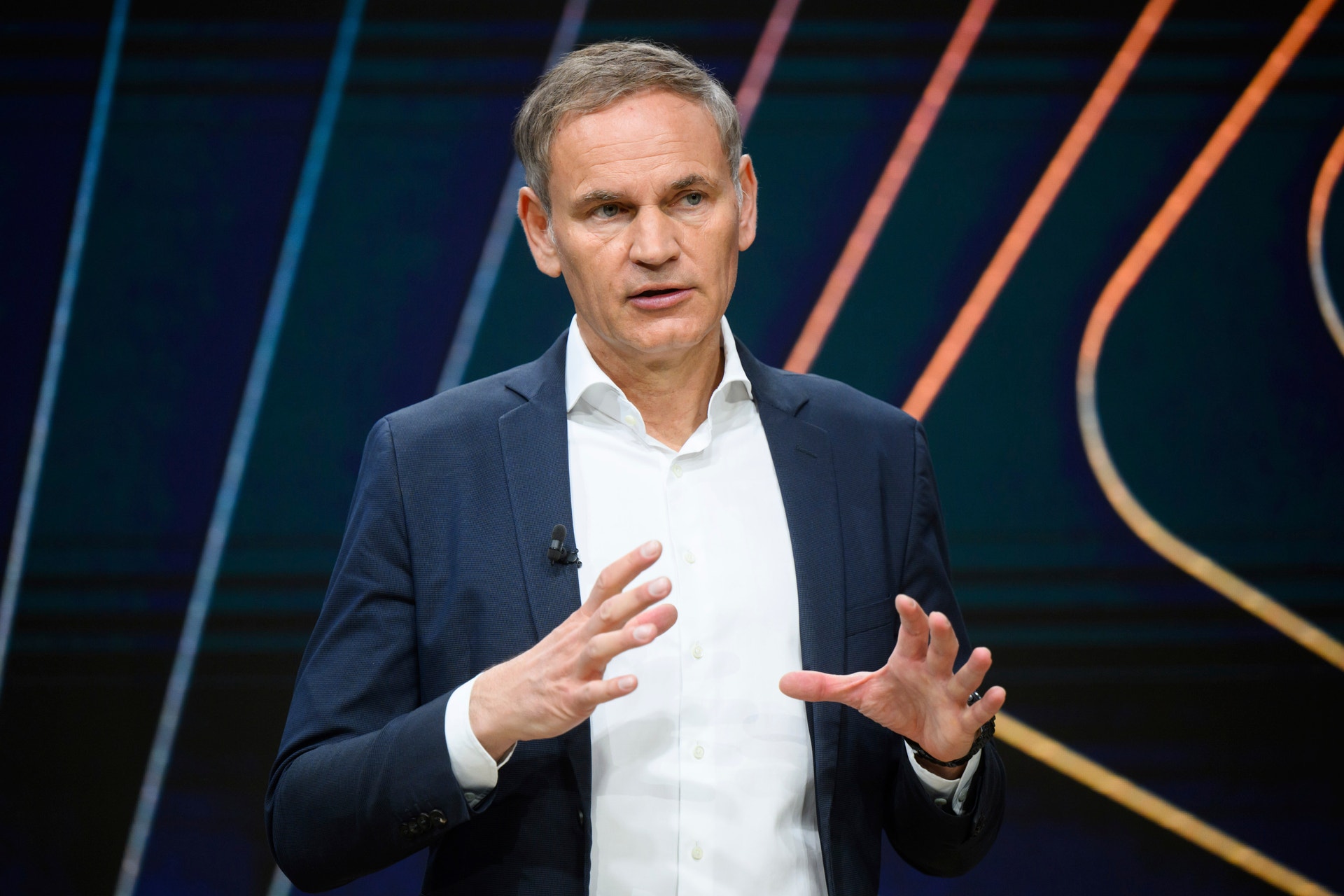 Man i mörk kostym och vit skjorta håller presentation på scen med färgad bakgrund