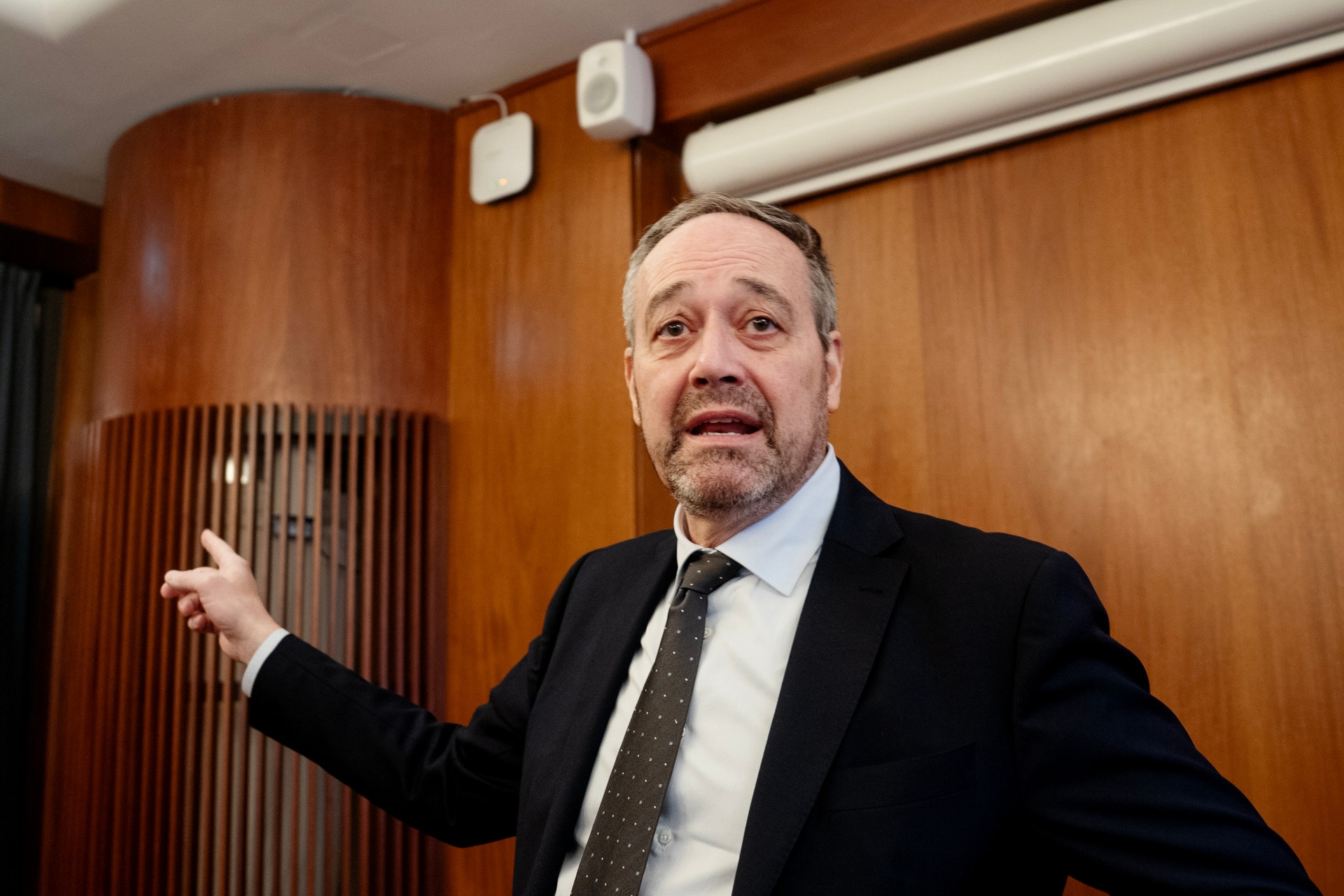 En man i kostym och slips talar framför en vägg med träpanel.