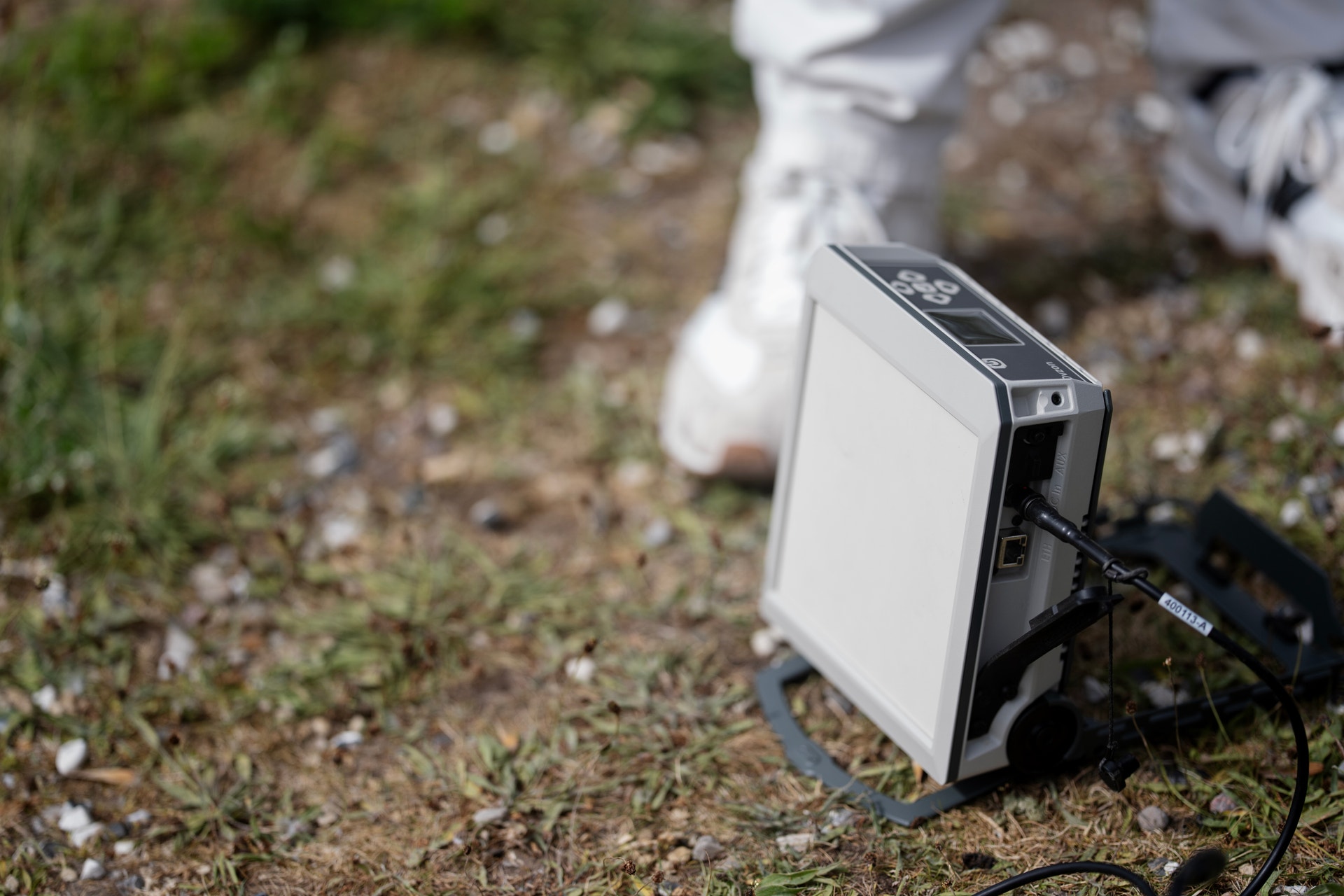 En vit och svart elektronisk enhet ligger på marken omgiven av gräs och stenar.