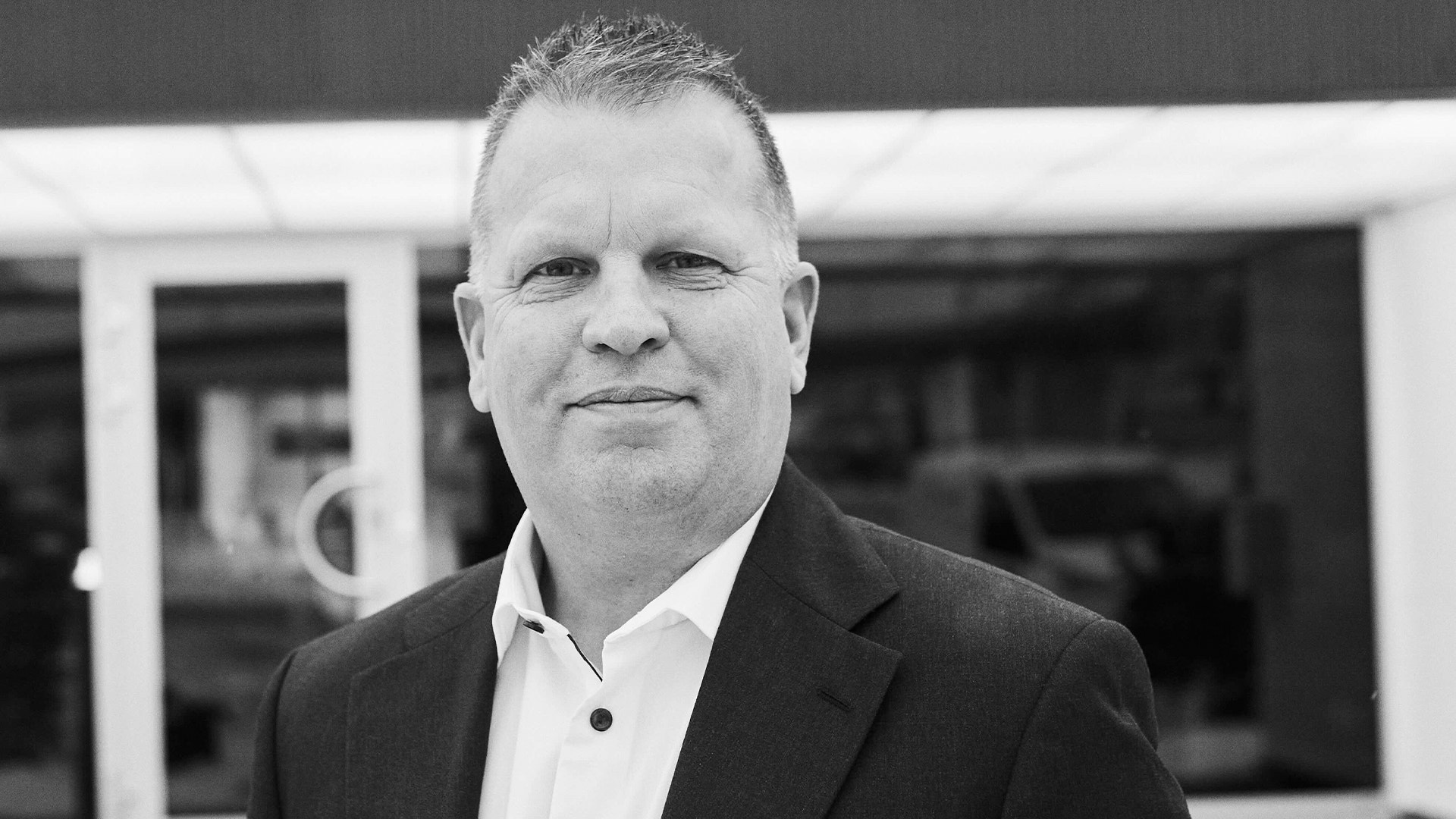 Svartvitt porträtt av en man i mörk kostym och vit skjorta framför en byggnad.