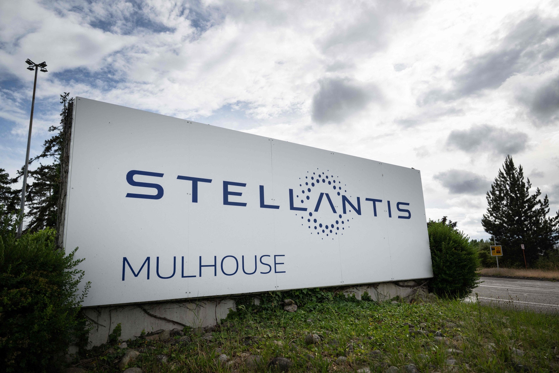 Stor vit skylt vid en väg med texten Stellantis och Mulhouse, omgiven av grönska under en molnig himmel.