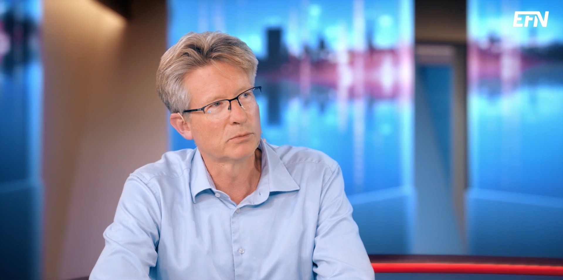 Man i ljusblå skjorta sitter framför en TV-studiobakgrund med blå och rosa neonljus.