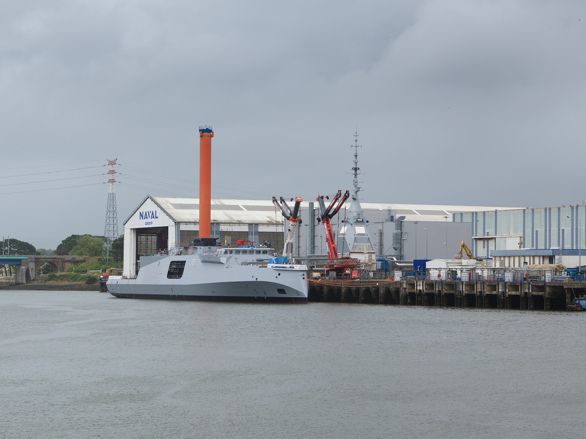 Ett modernt fartyg med vit skrov och (orange skorsten bakom) dockat vid en industriell kaj vid en flod.