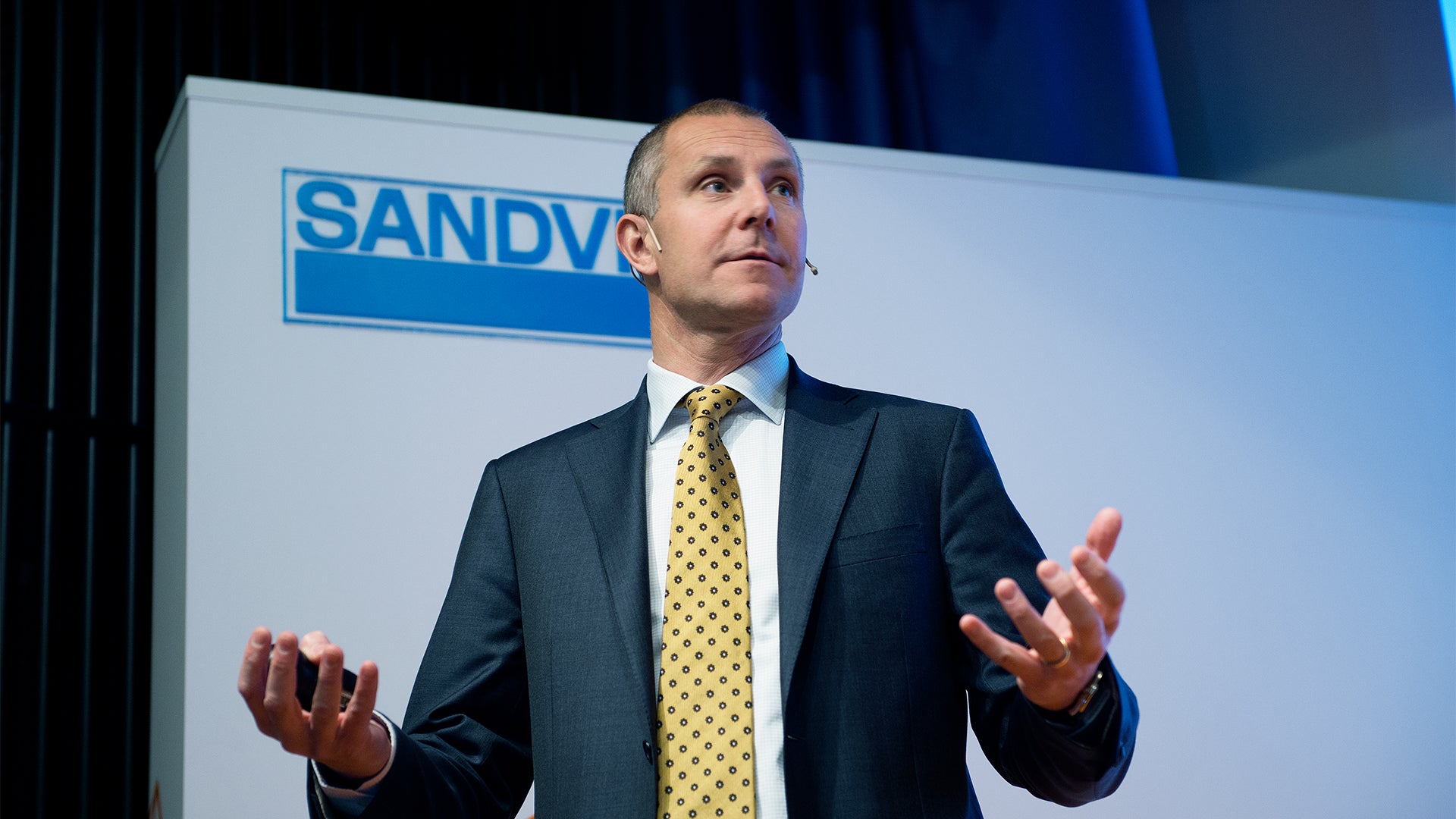 Man i mörkblå kostym och gul slips presenterar på scen framför blå bakgrund med Sandy-logotyp.