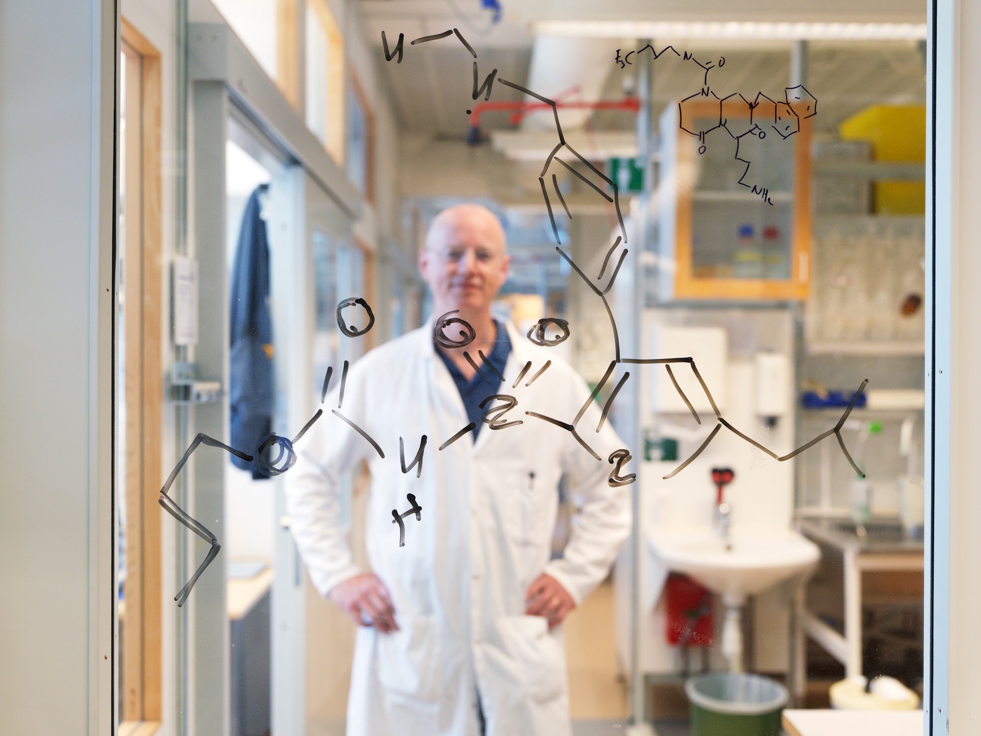 Forskare i vit laboratoriekittel står i ett laboratorium med ritade kemiska molekylstrukturer på glaset framför honom.