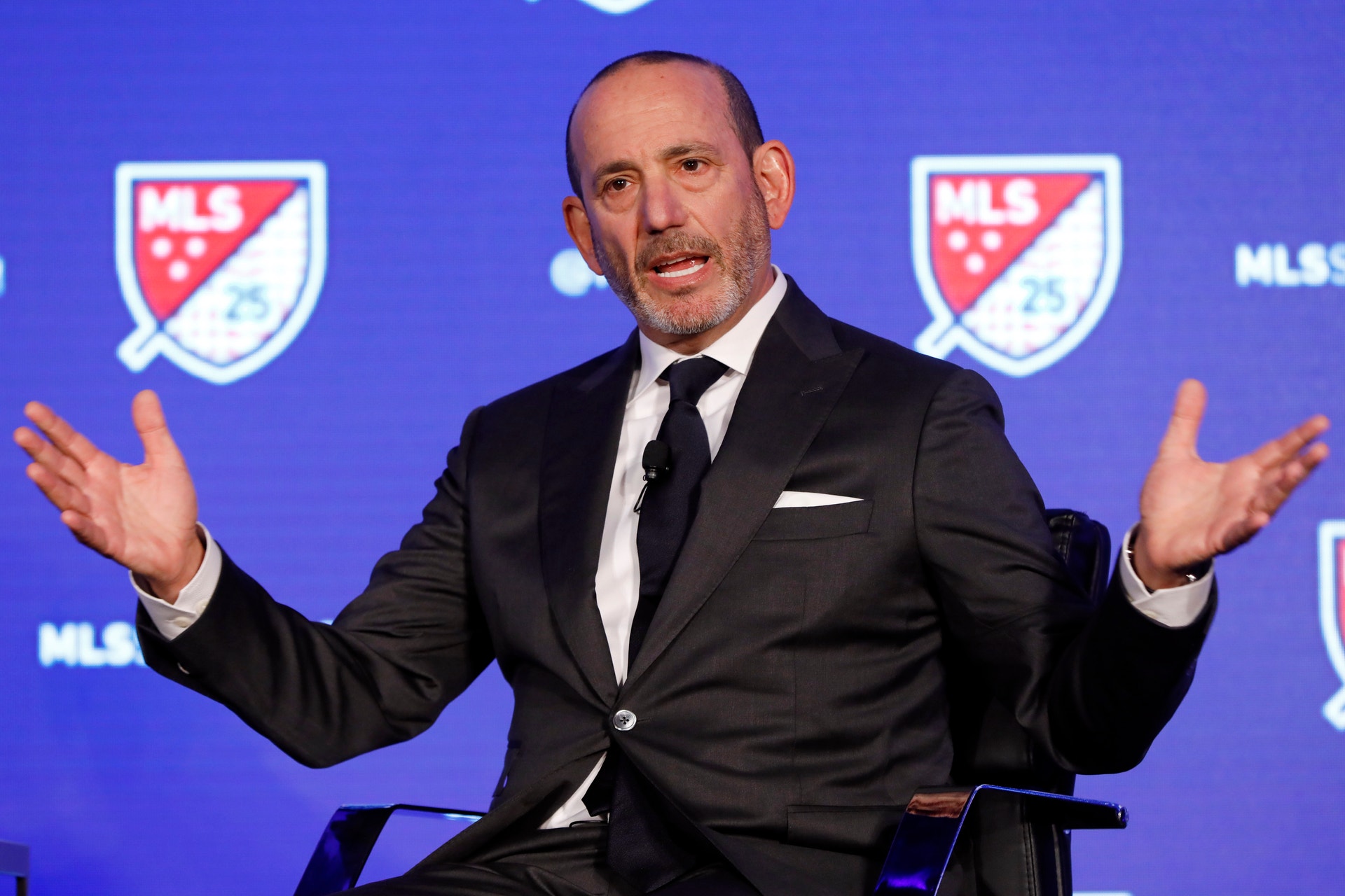 En man i svart kostym och vit skjorta talar vid ett podium framför en blå bakgrund med MLS-logotyper.