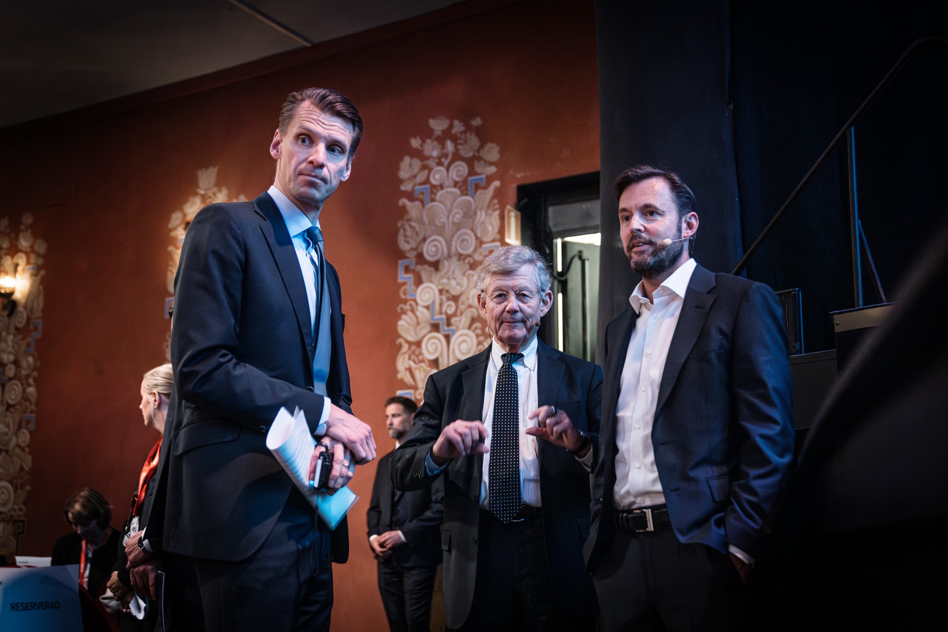 Tre män i kostym står tillsammans i en festlig lokal med dekorerade väggar. Två av dem tittar mot något utanför bild, och mannen i mitten gestikulerar medan han pratar.